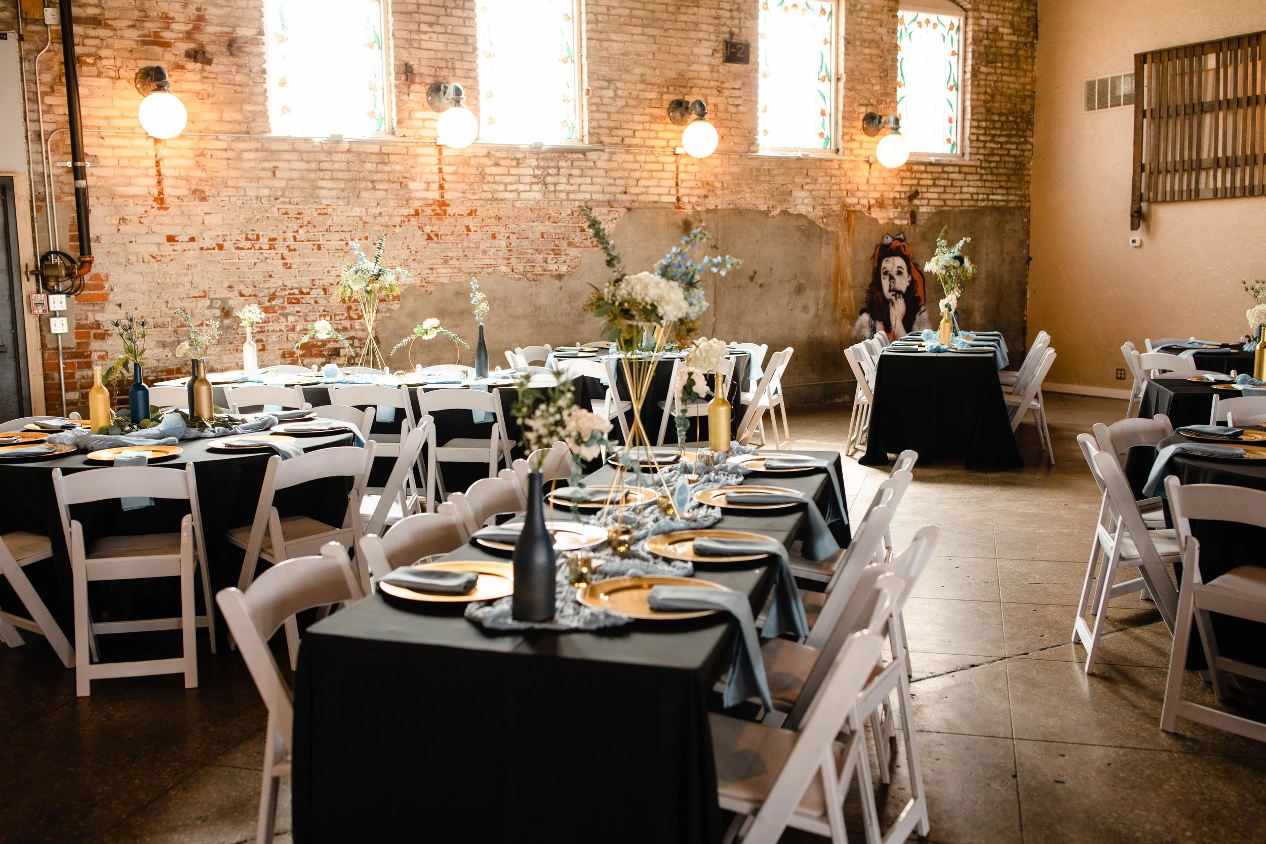 Event space decorated with black tablecloths, white chairs, and vases with white and pink flowers, set for a banquet with plates, napkins, and utensils. Exposed brick wall with stained glass windows and a side wall with a portrait painting.