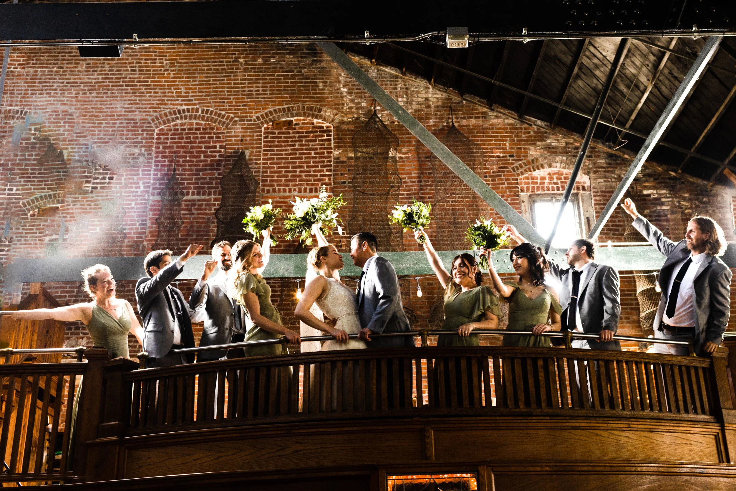 A wedding celebration with the bride and groom kissing on a wooden balcony, surrounded by their bridal party holding bouquets of flowers, inside a rustic brick venue with high ceilings.