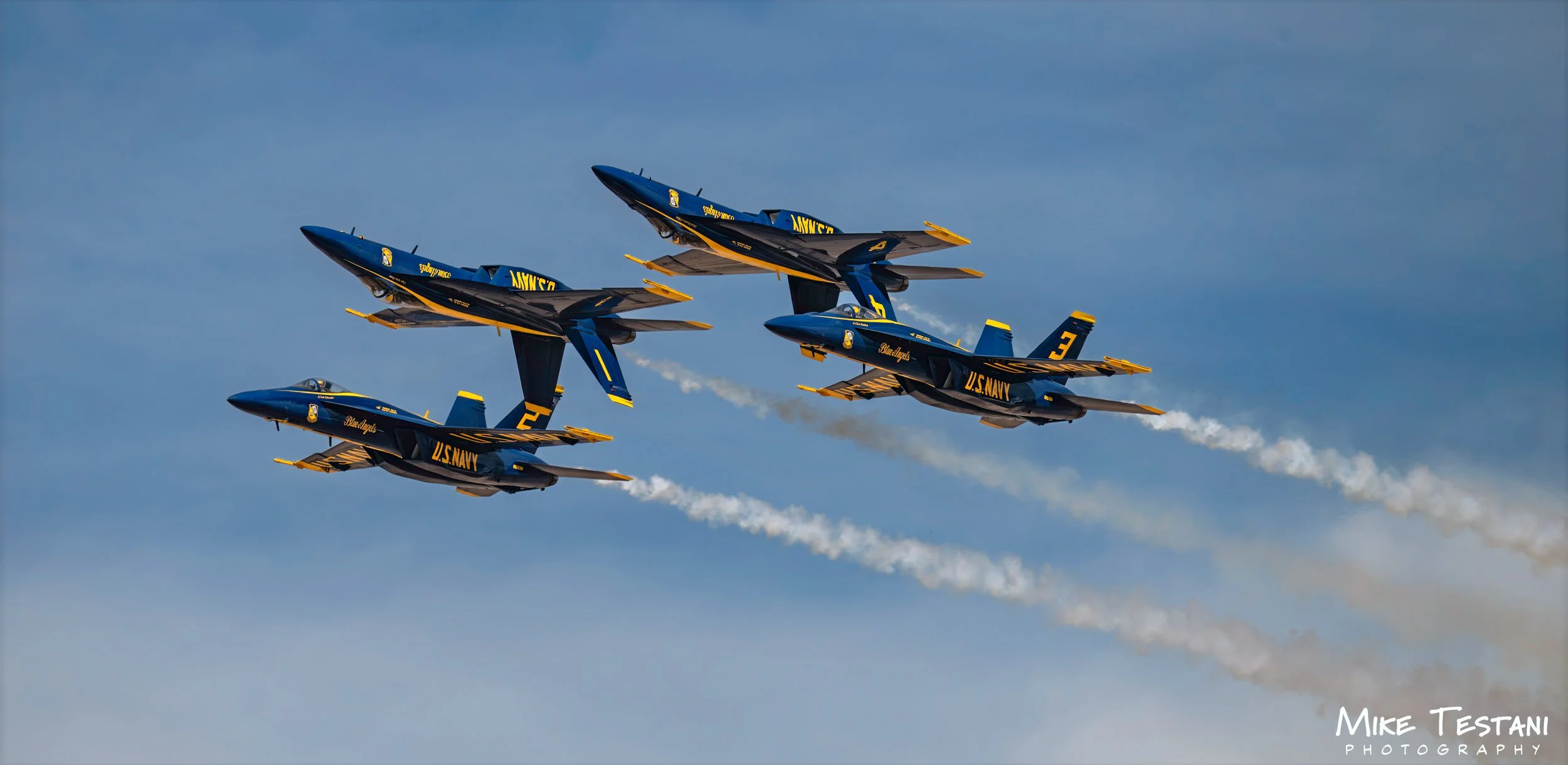 A formation of five US Navy Blue Angels aircraft flying in close proximity, emitting white smoke trails against a blue sky.