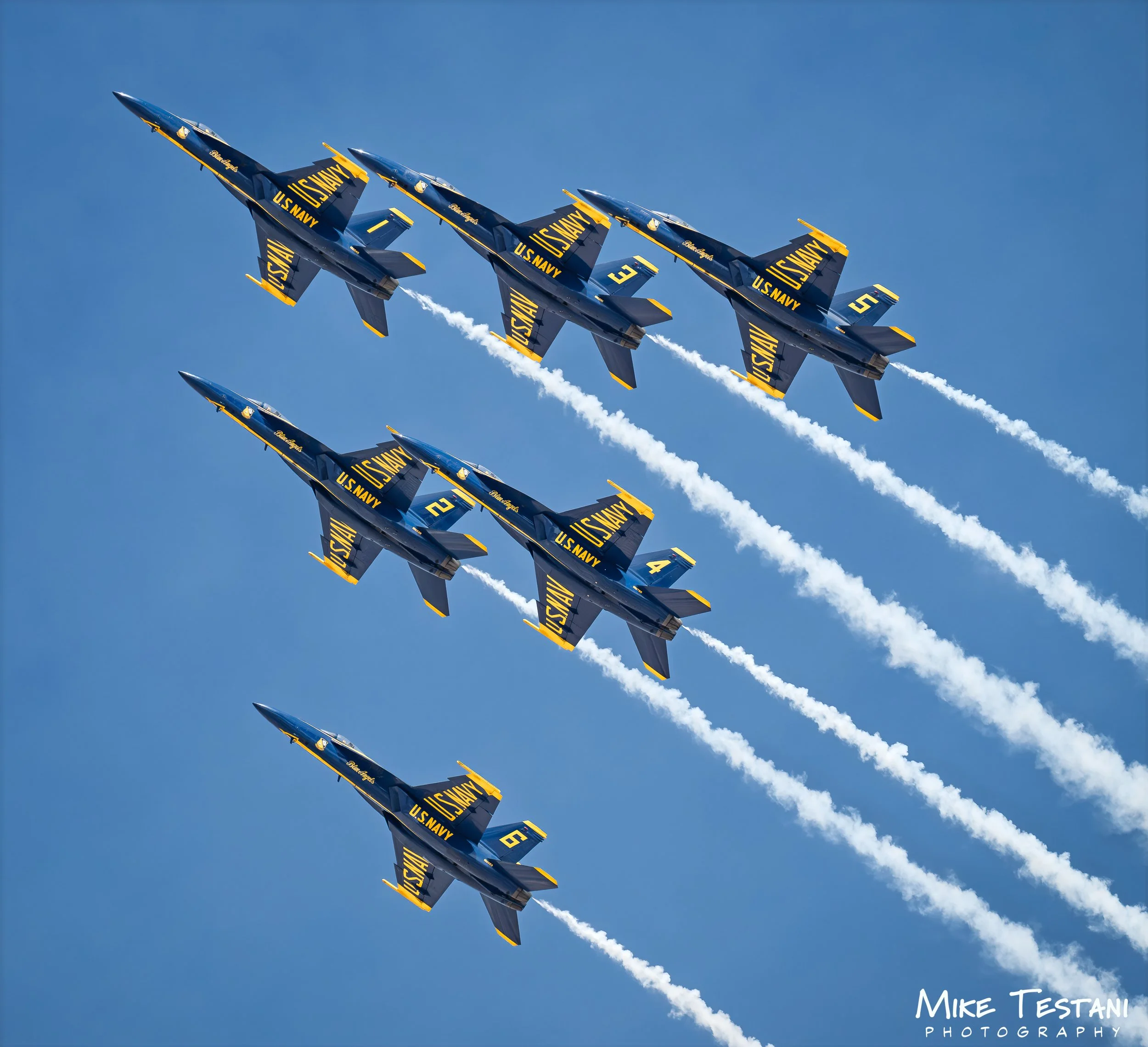 A group of blue U.S. Navy fighter jets flying in close formation against a clear blue sky, leaving white smoke trails behind them.