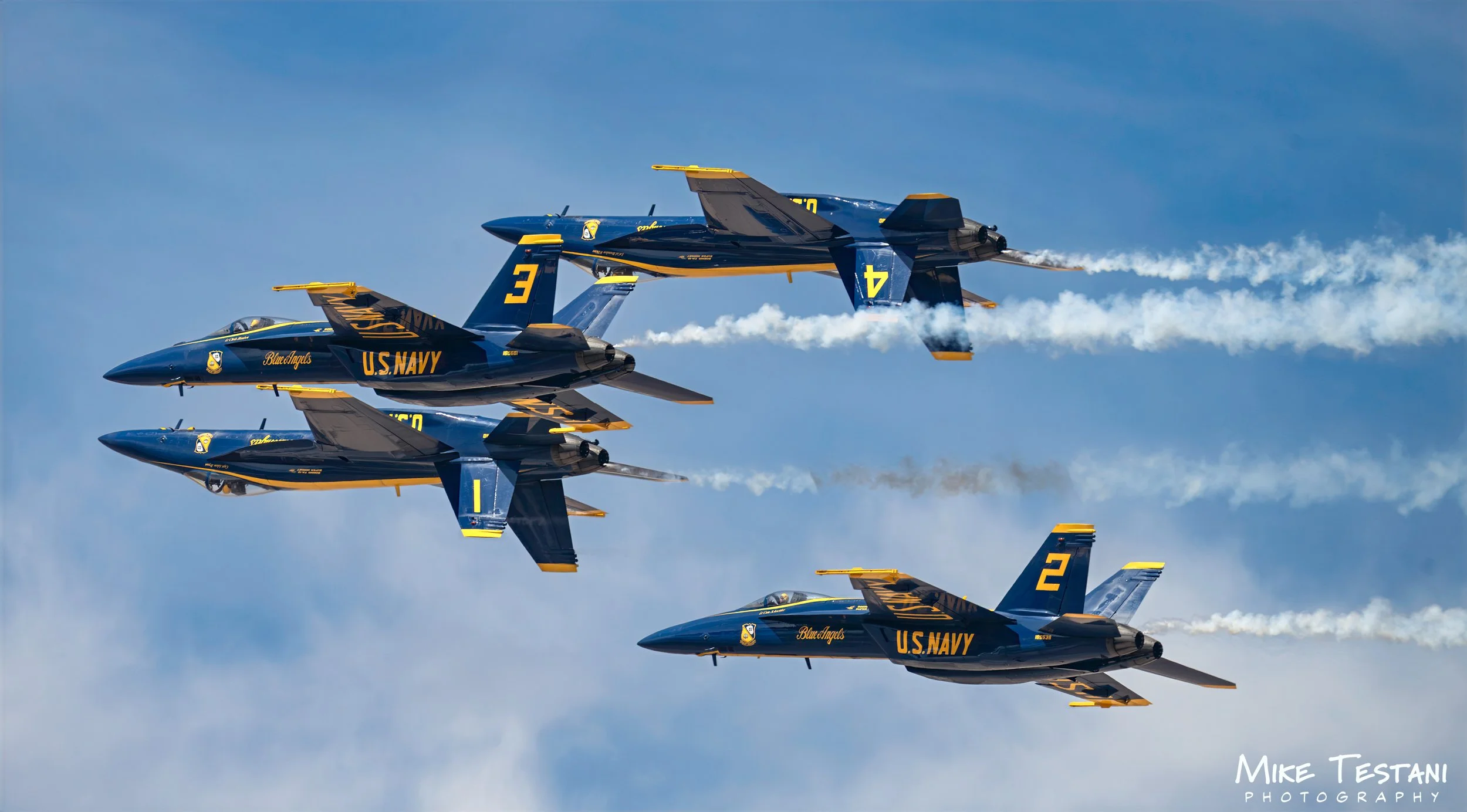 Aerial display team of four U.S. Navy Blue Angels fighter jets flying in close formation through the sky, leaving white smoke trails behind them.