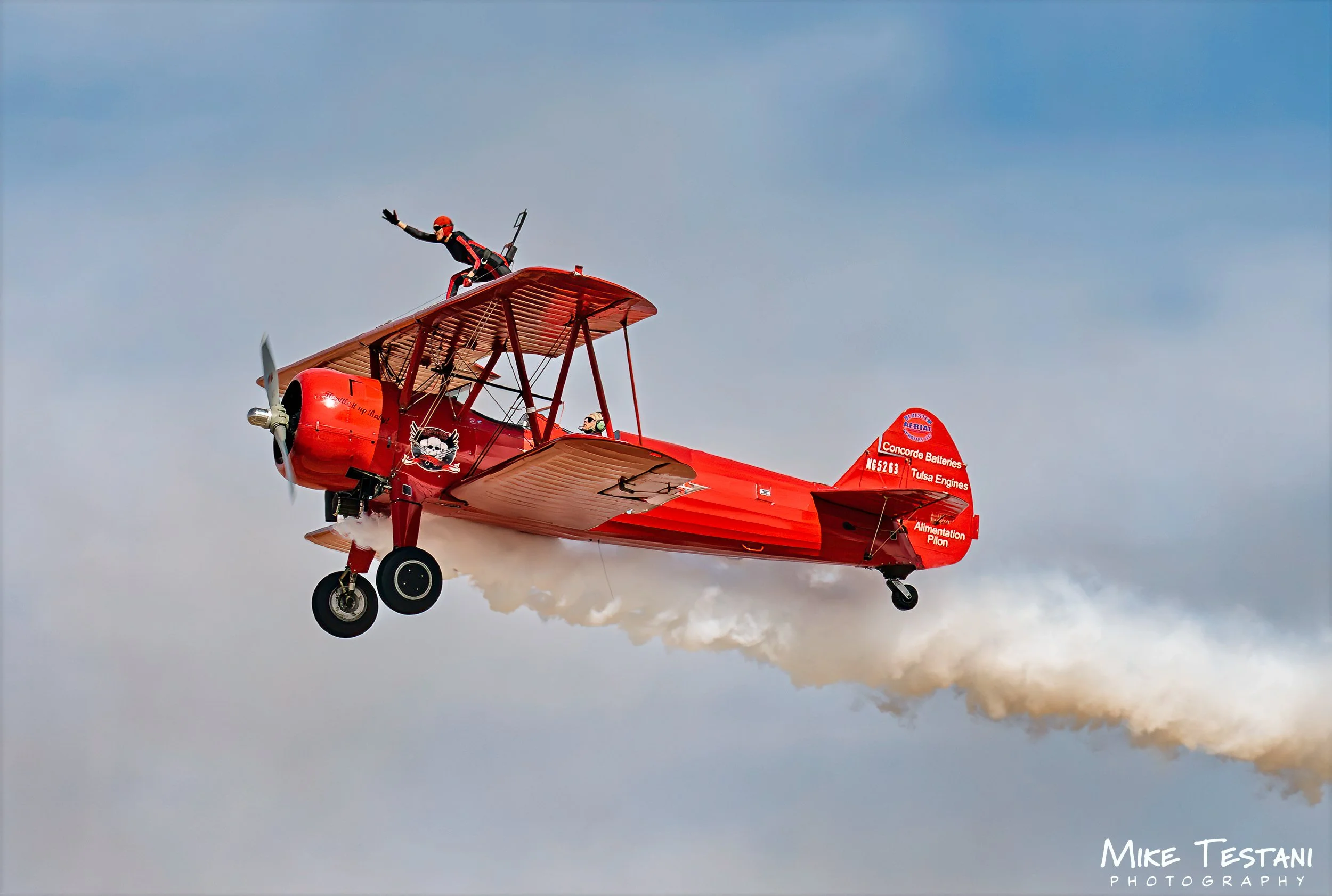 A red biplane airplane flying in the sky with a pilot and a passenger on board. The airplane has a skull logo on its side and smoke trails behind it.