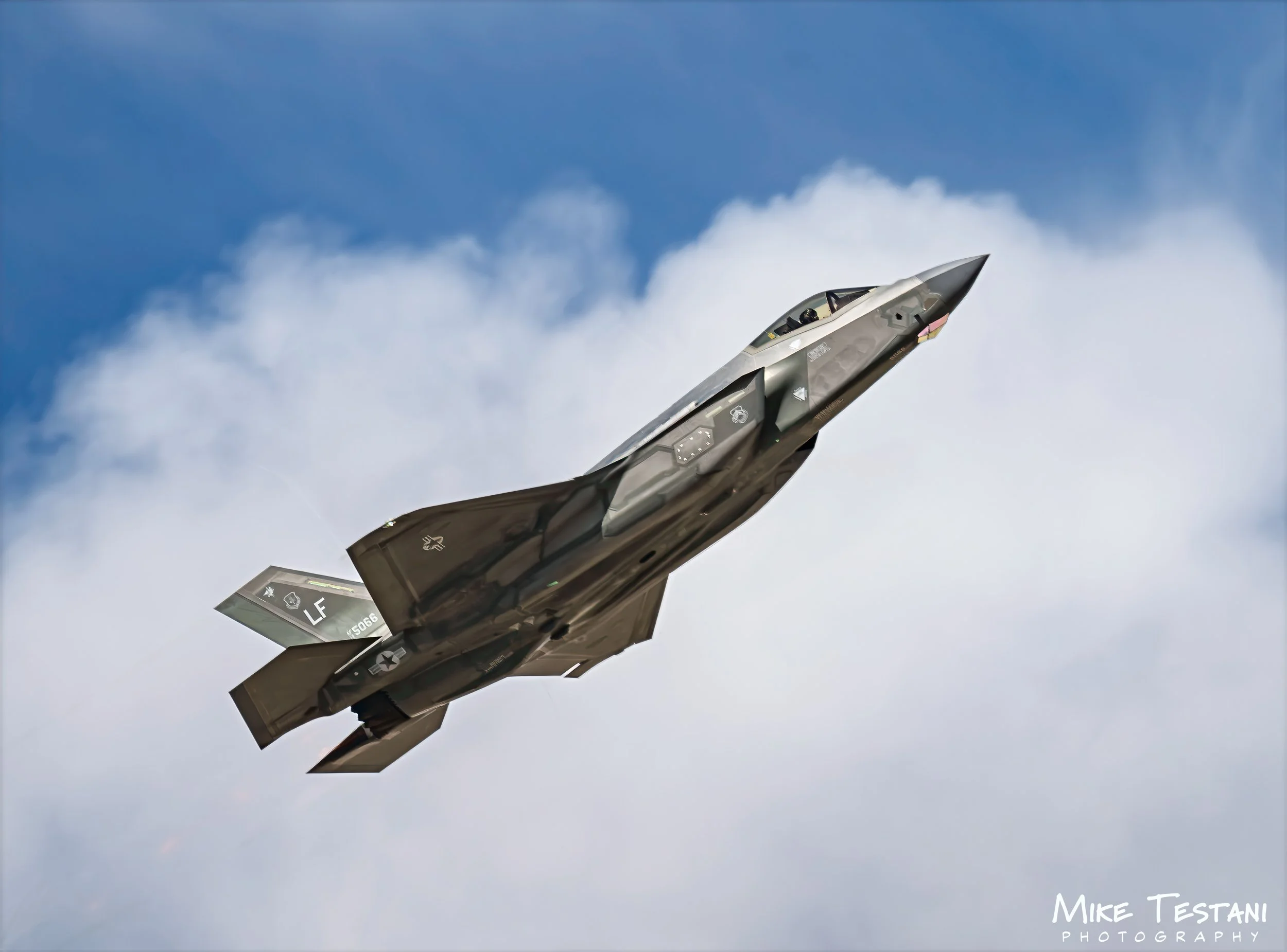 A military fighter jet in flight against a blue sky with white clouds.