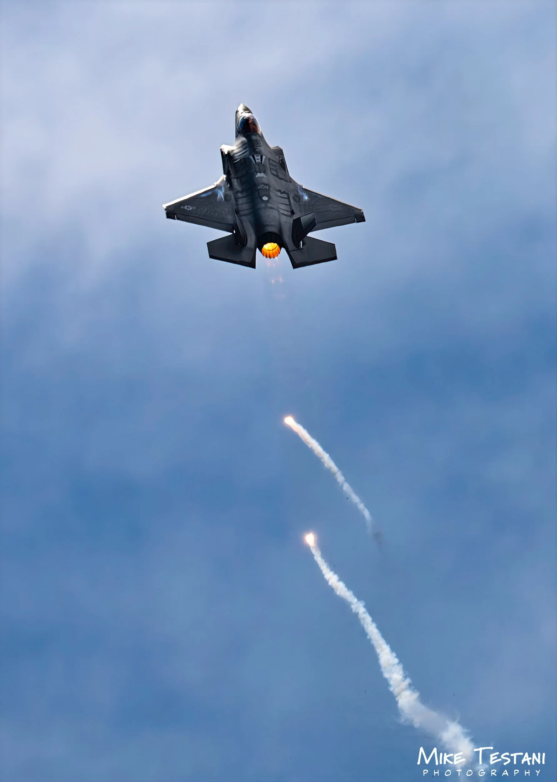 An F-22 fighter jet flying upwards through a blue sky, releasing a fiery afterburner and producing a trail of smoke.