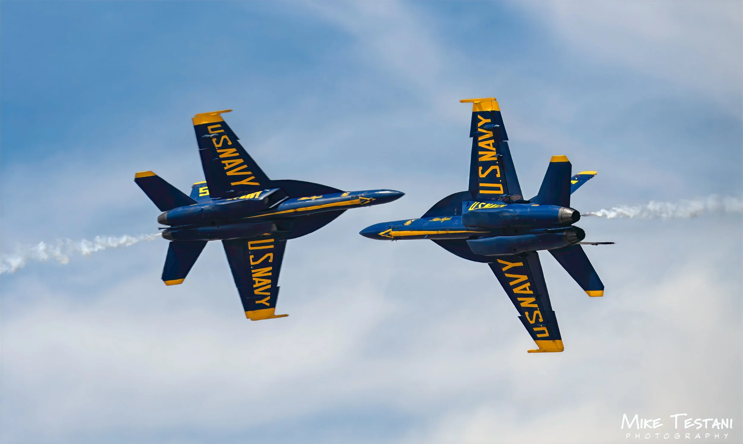 Two blue fighter jets with yellow accents, marked with 'U.S. NAVY', flying in close formation against a cloudy sky, leaving white smoke trails.