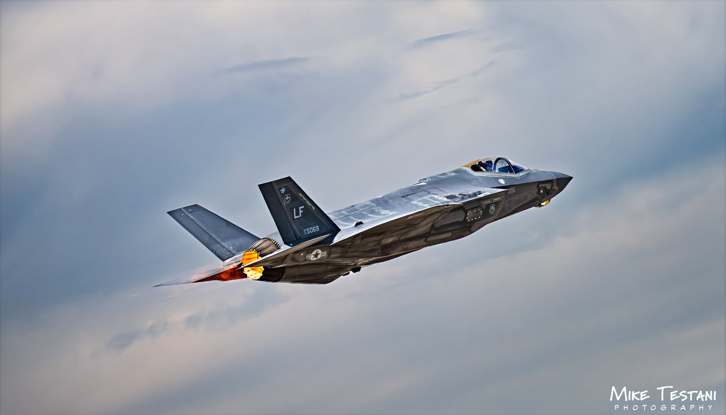 A fighter jet flying through the cloudy sky with afterburners lit.
