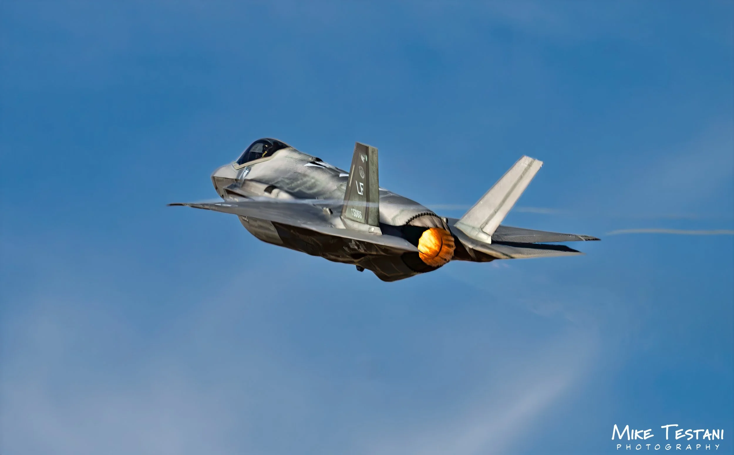 A fighter jet airplane in flight against a clear blue sky, with afterburner flames visible at the jet's exhaust.