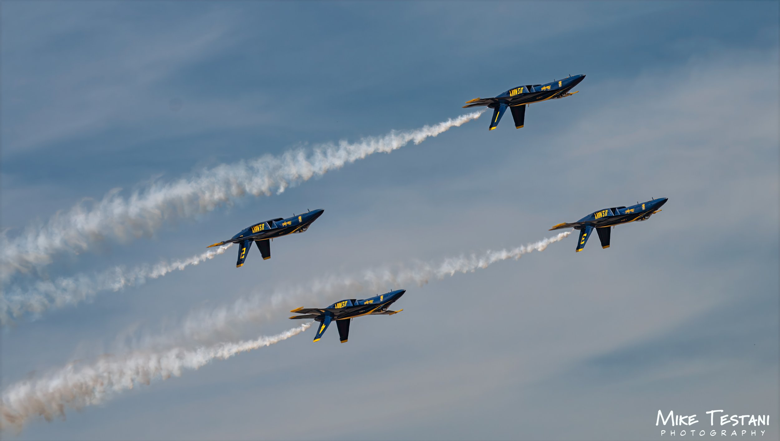 Five blue and yellow jets flying in formation, leaving white smoke trails against a partly cloudy sky during an air show.