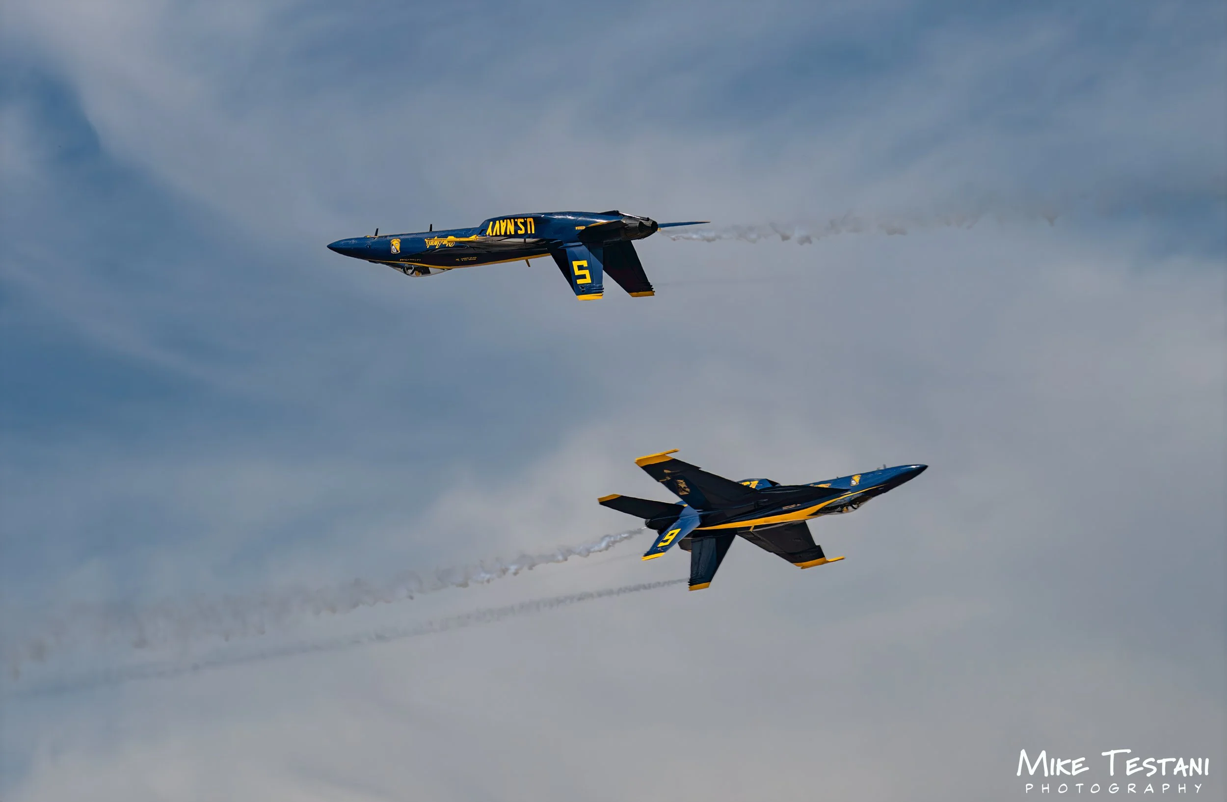 Two blue and yellow fighter jets perform an aerial stunt in the sky, leaving smoke trails behind.