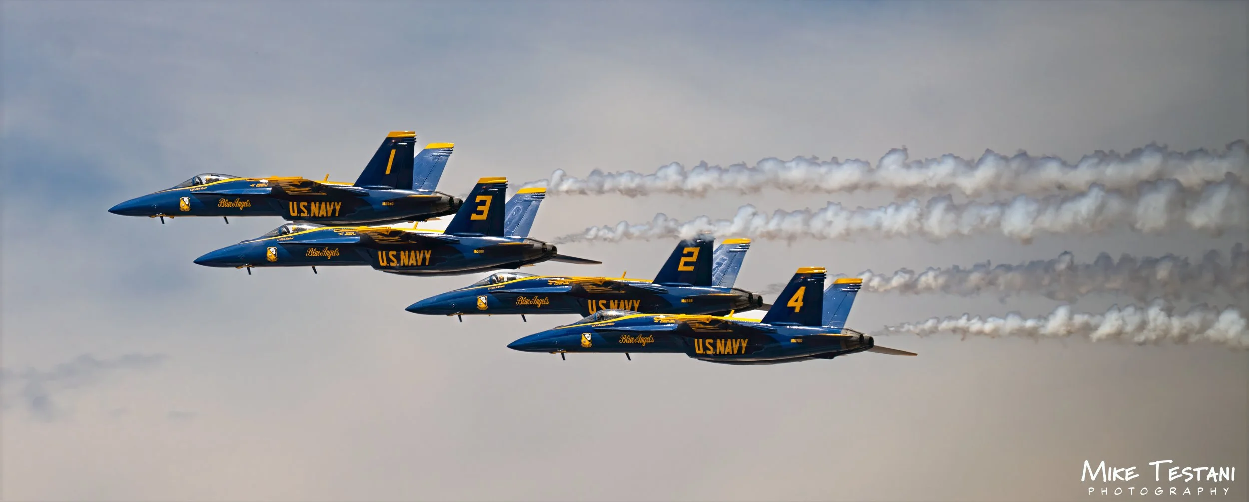Four U.S. Navy Blue Angels fighter jets flying in close formation, leaving white smoke trails against a cloudy sky.