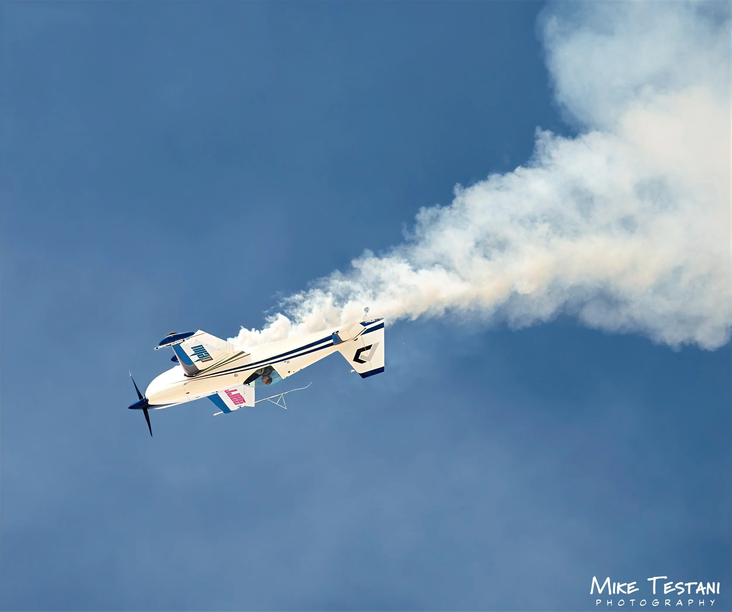 A white and blue aerobatic airplane flying through a clear sky with some clouds, leaving a trail of white smoke behind.