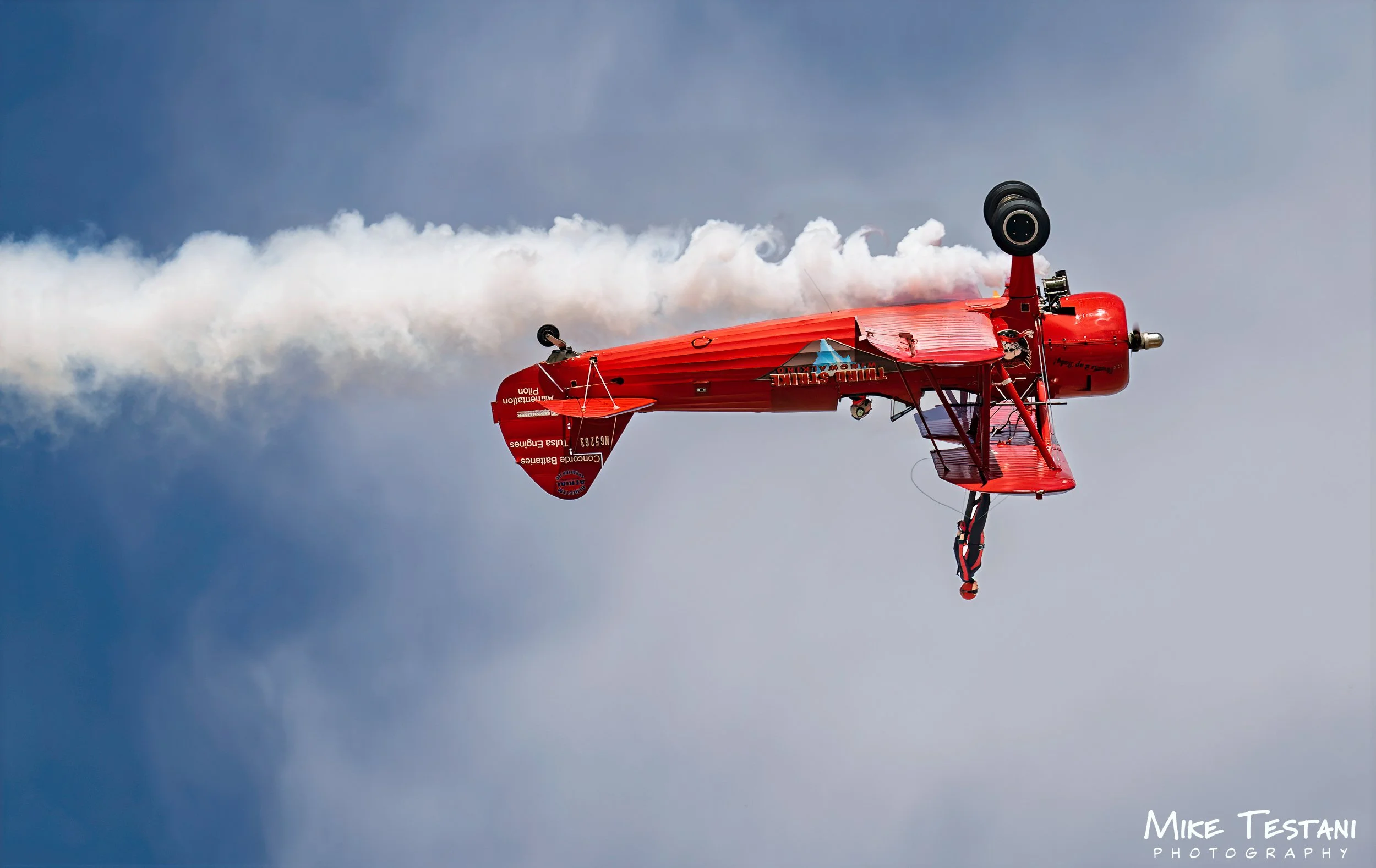 A red stunt plane performing a corkscrew maneuver in the sky, emitting white smoke trail.