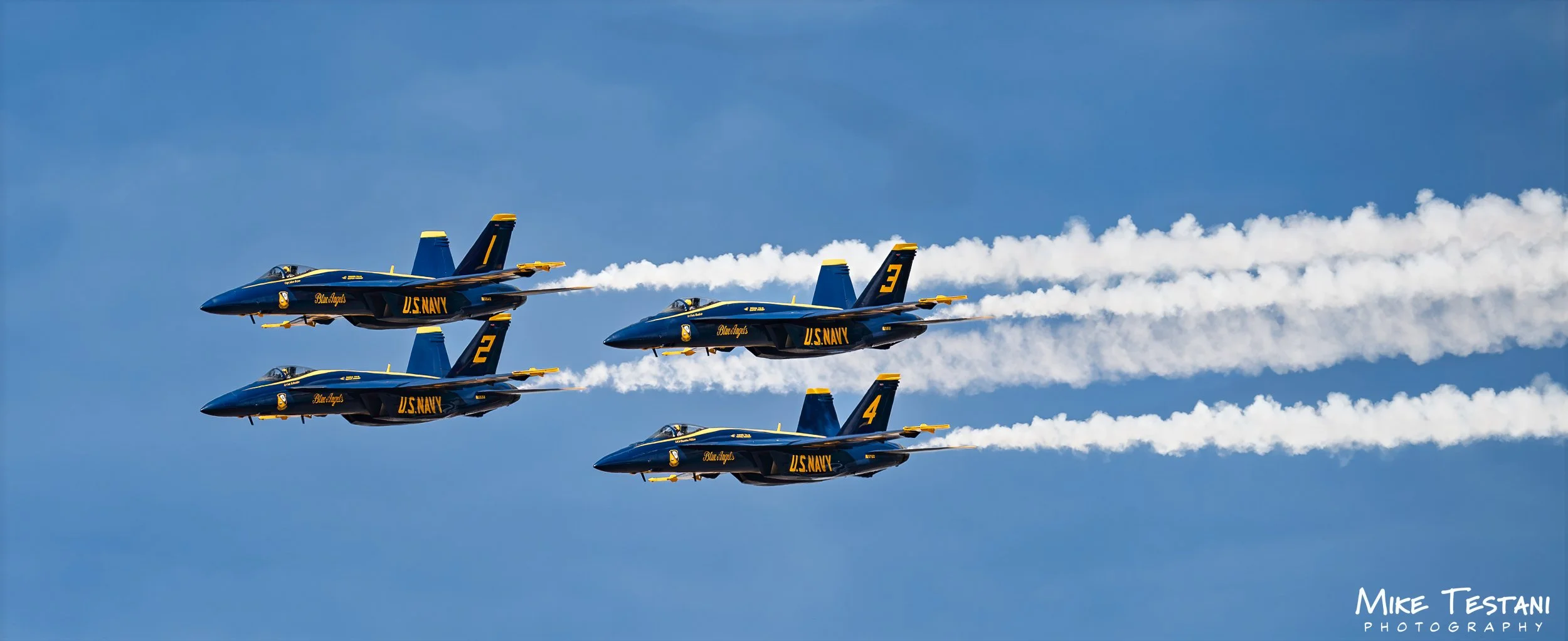 Four U.S. Navy Blue Angels jet fighters flying in tight formation with trails of white smoke against a blue sky.