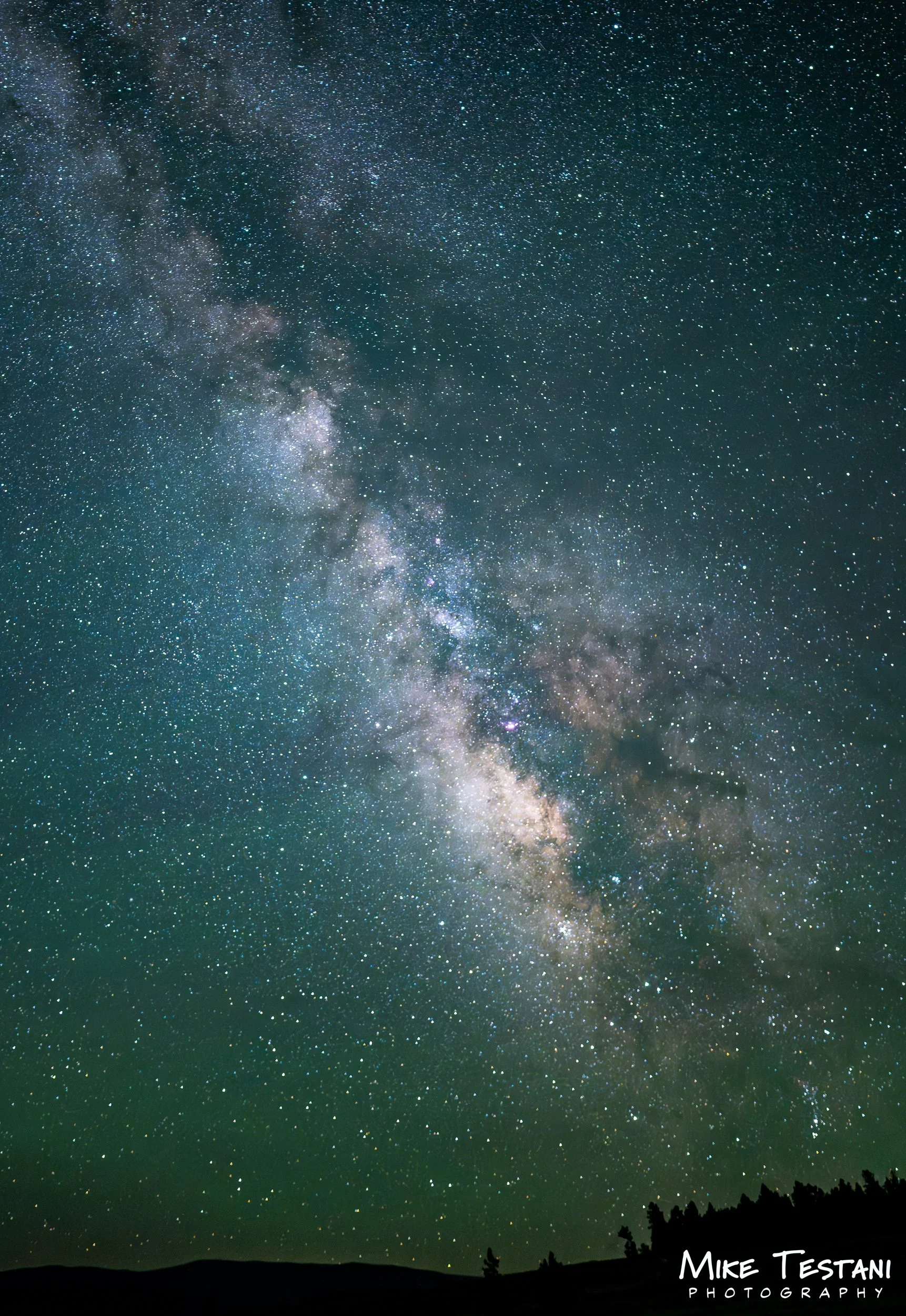 A starry night sky with the Milky Way galaxy visible, stretching across the horizon over a silhouette of trees and rolling hills.