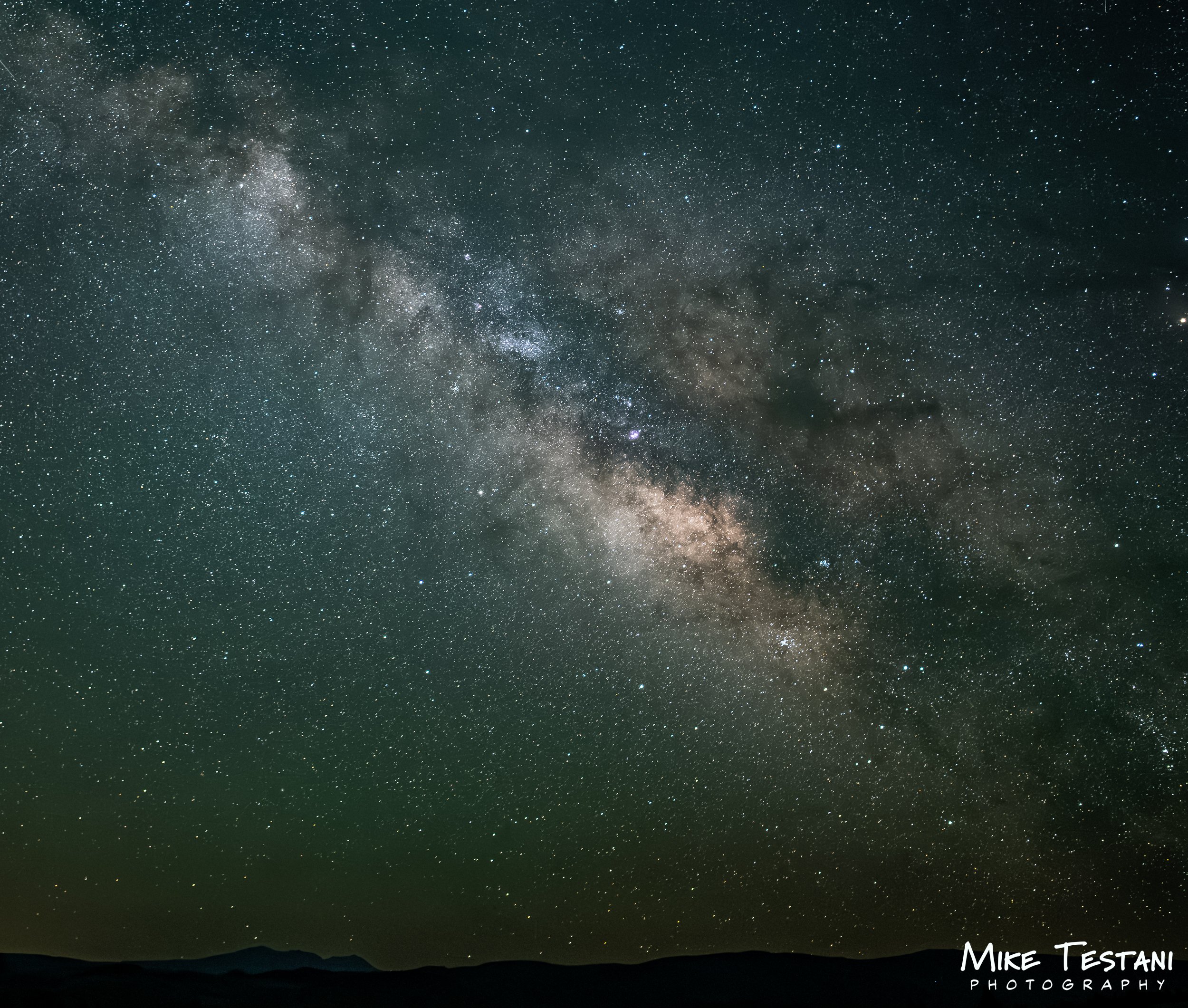 View of the night sky filled with many stars and the Milky Way galaxy, with a dark silhouette of mountains or hills at the bottom.