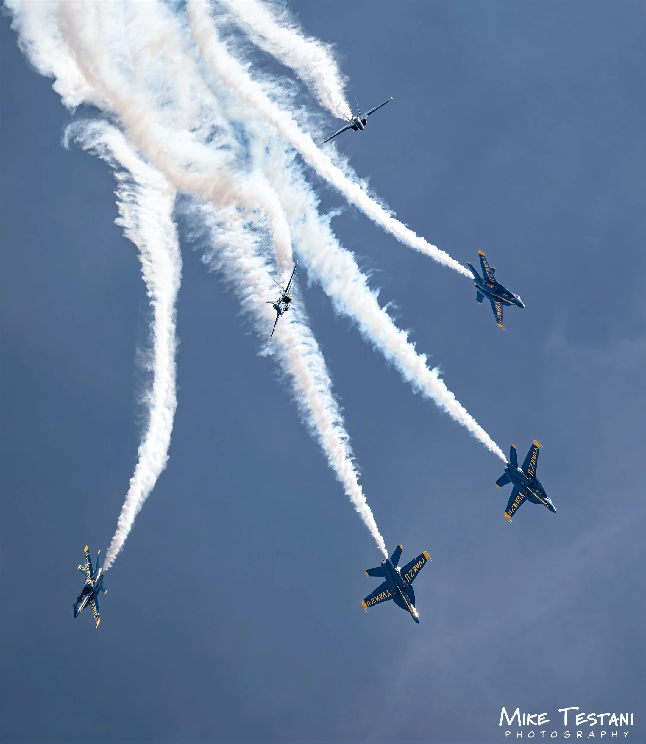 Four aerobatic aircraft performing a synchronized stunt in the sky, leaving white smoke trails behind them against a clear blue sky.