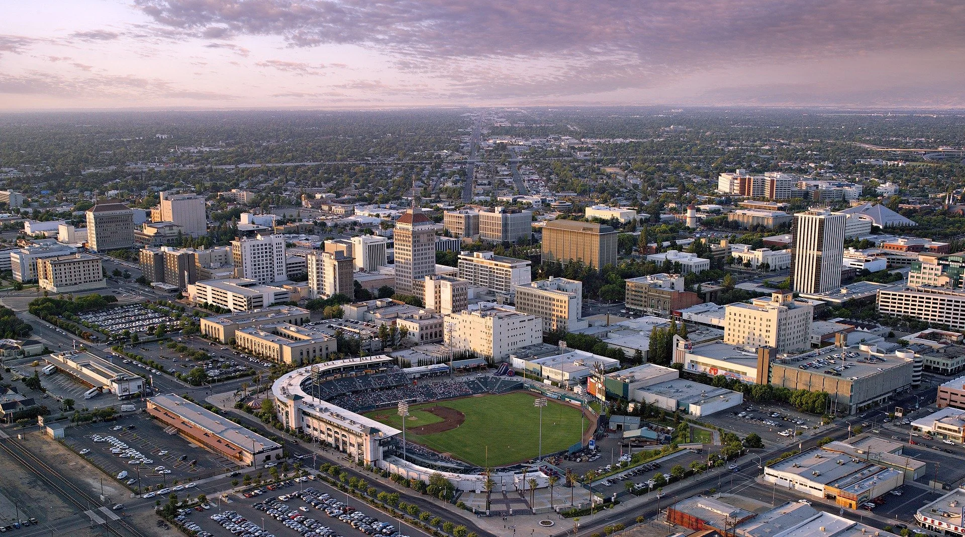 Aerial view of a cityscape at dusk featuring tall buildings, a baseball stadium with a green field, parking lots, and streets extending into the distance under a cloudy sky.
