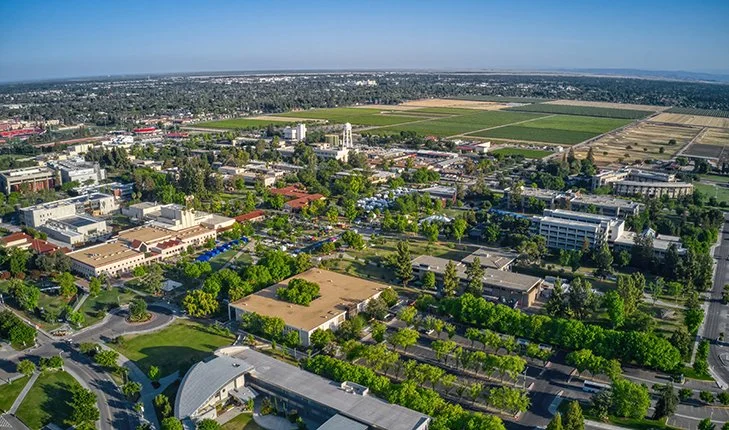 An aerial view of a city with a mix of buildings, roads, and green spaces, including agricultural fields in the distance.