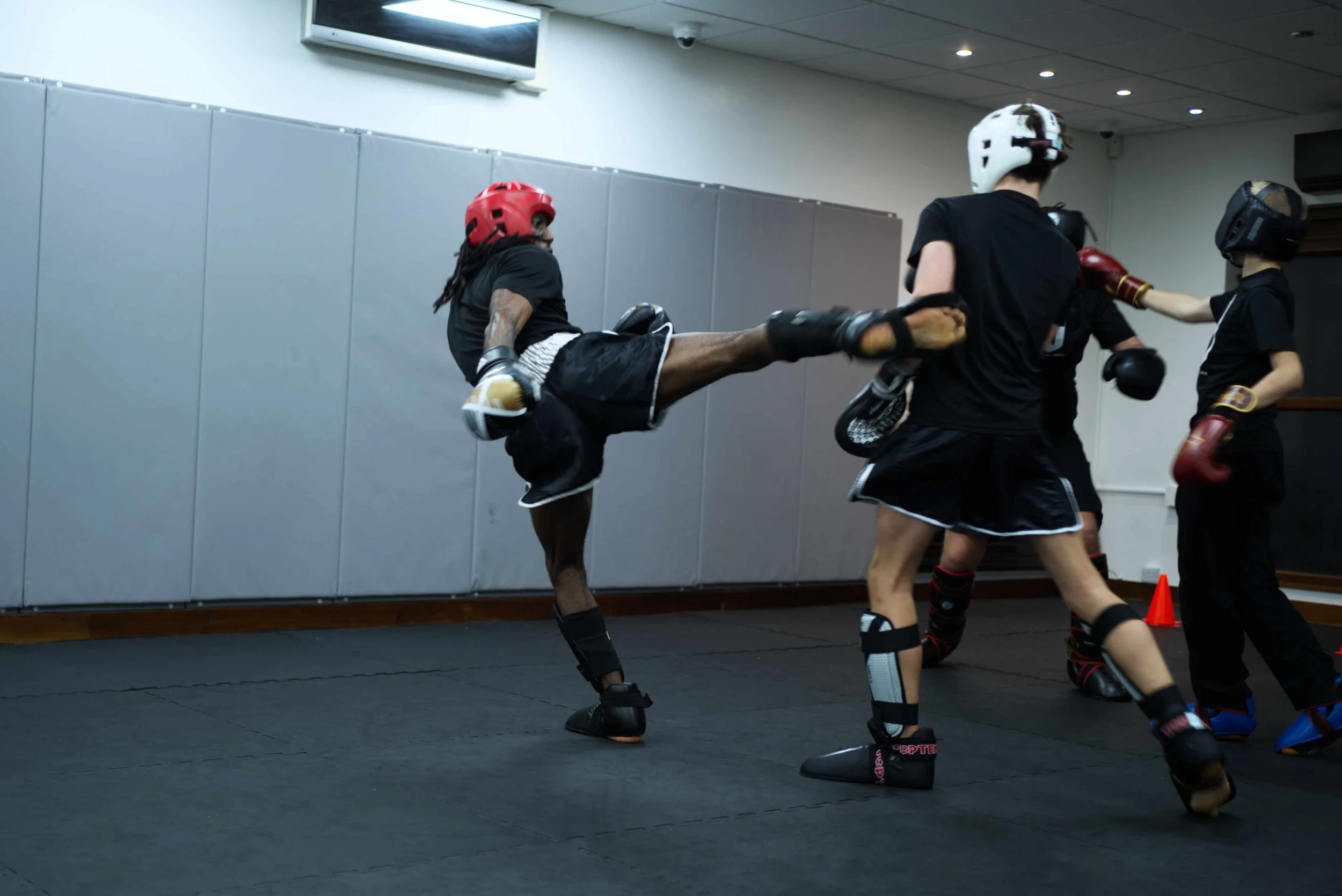Martial artists practicing kickboxing in a training room, wearing protective gear and black uniforms, with one executing a high kick.