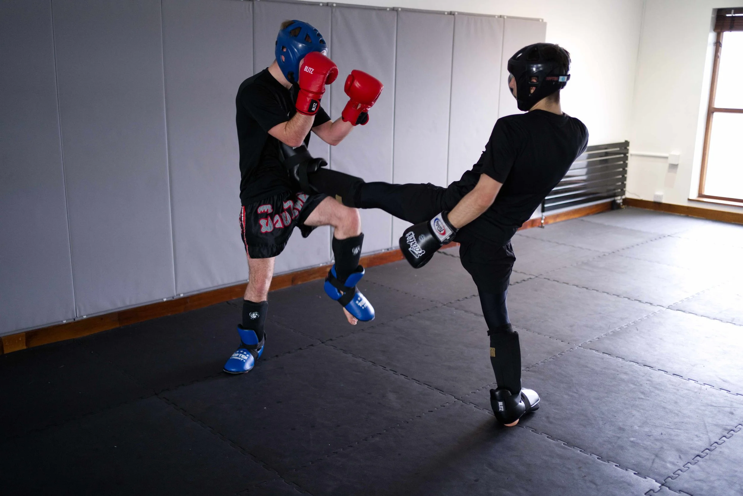 Two martial artists wearing protective helmets and gloves practice kickboxing in a gym, with one executing a high kick towards the other's head.