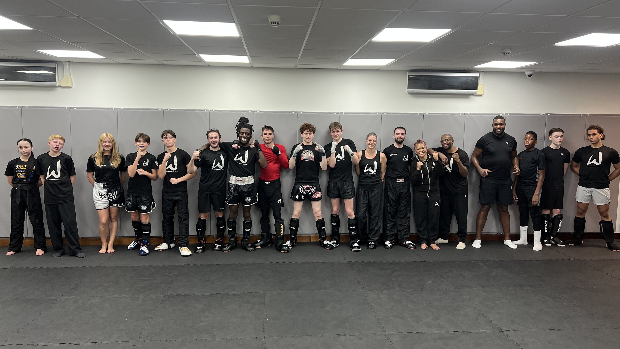 Group of mixed gender martial artists and coaches in a gym, wearing training gear and standing in a line, some in fighting stances.
