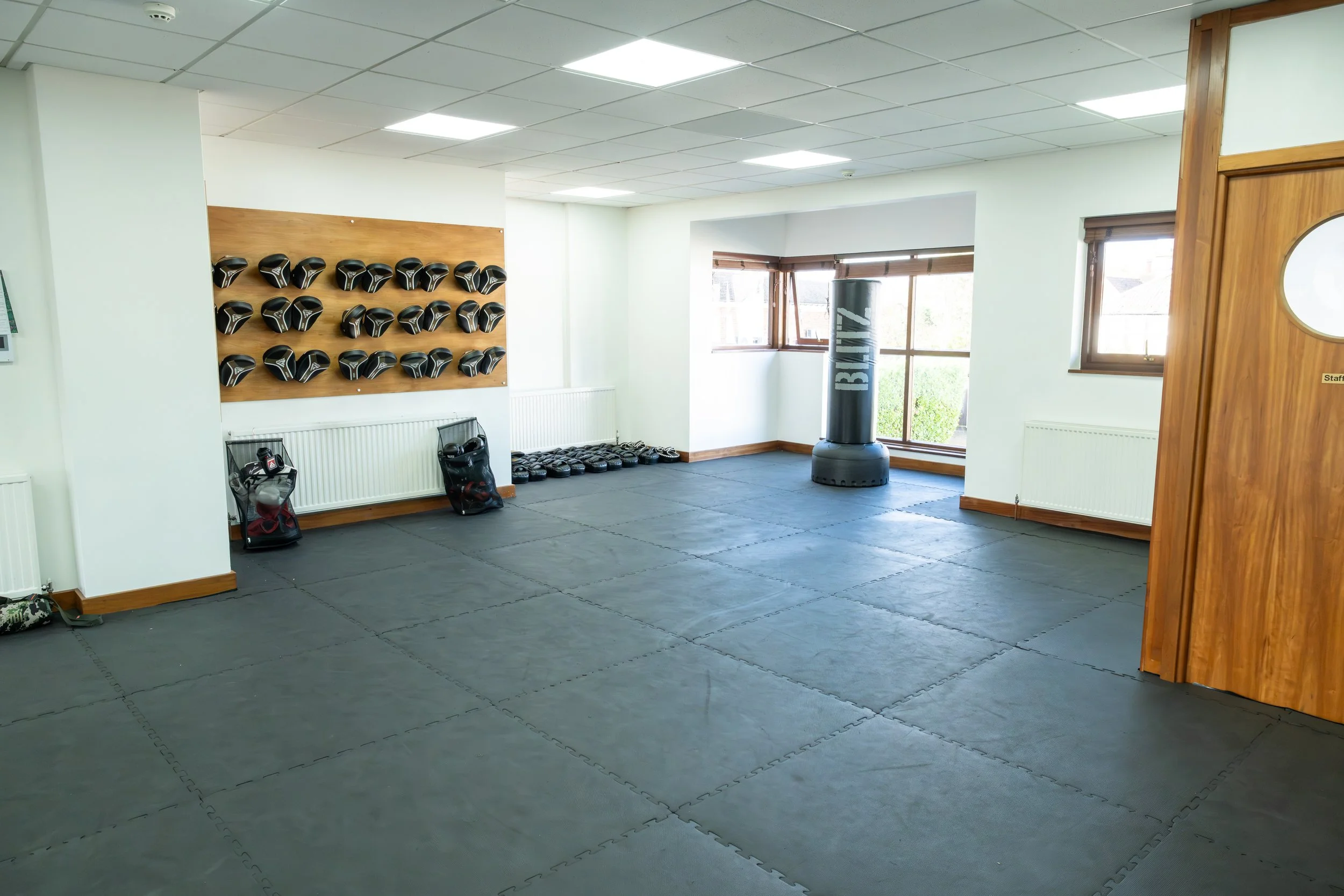 Empty martial arts training room with black mats on the floor, a wall-mounted rack of black and silver helmets, stacked black weight plates, a large black punching bag labeled BLITZ, and windows with natural light.