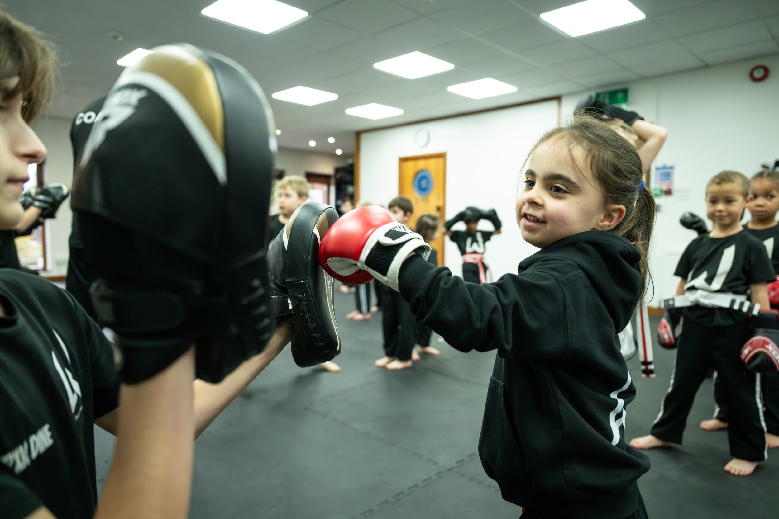 Young girl practicing boxing in a martial arts class, wearing boxing gloves, with other children in uniforms in the background.