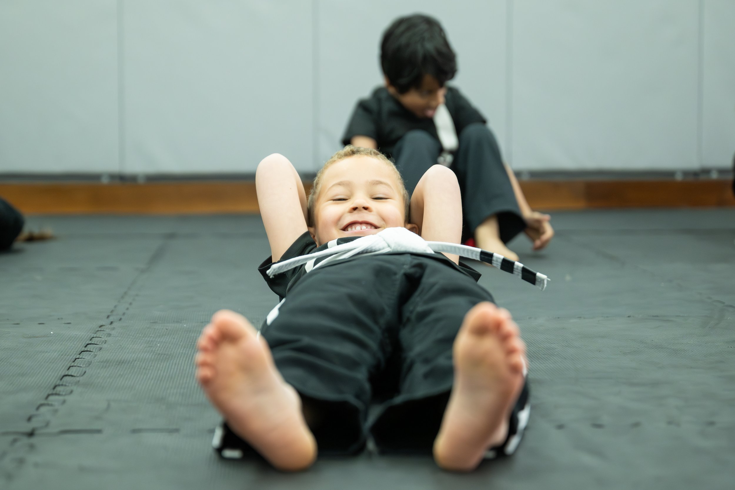 A girl lying on her back on a martial arts mat, smiling with her hands behind her head, while a boy sits in the background.