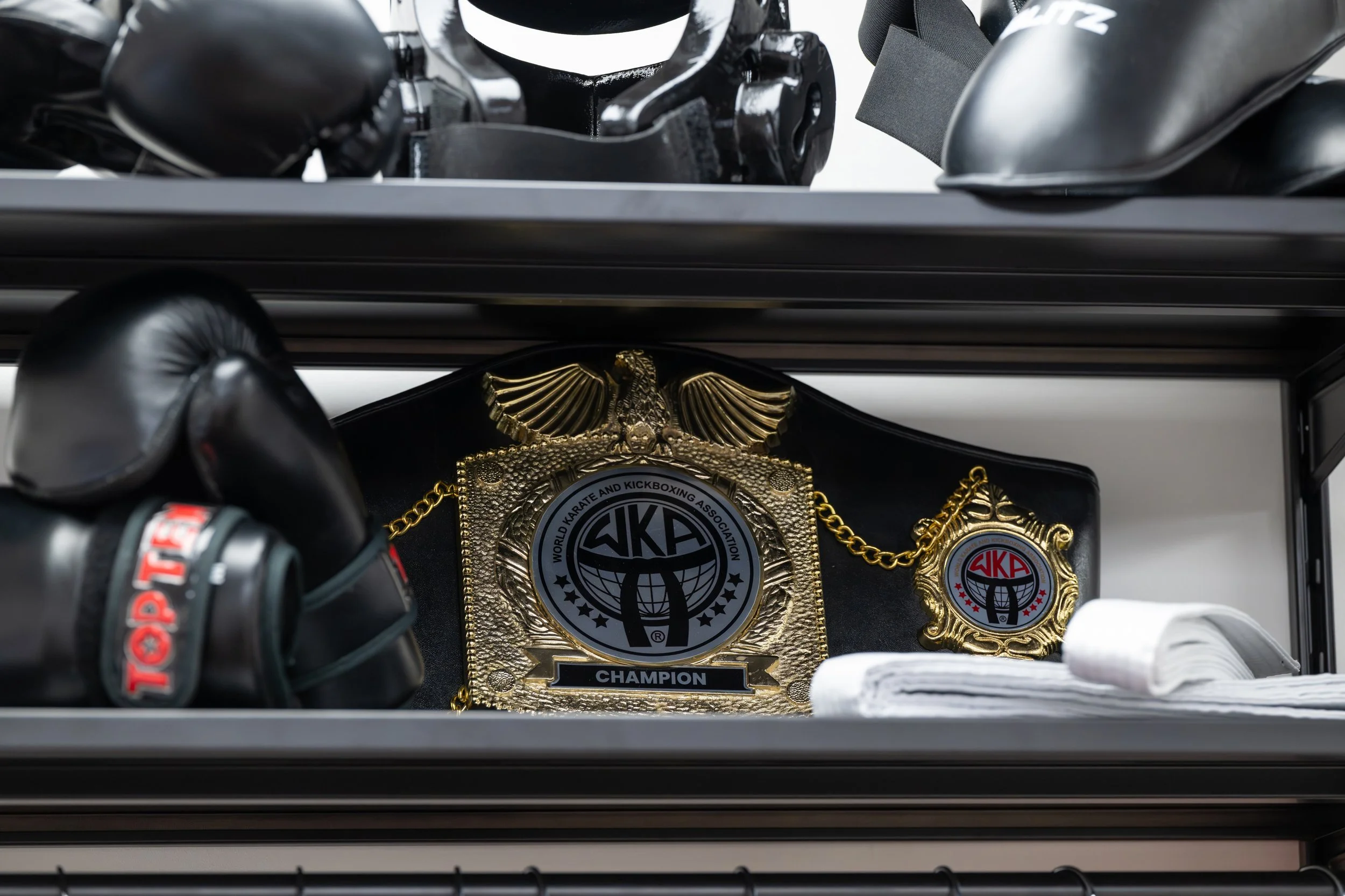 A wrestling championship belt with gold accents, the logo of World Karate and Kickboxing Association, and a badge with the same logo, placed on a black surface next to white towels and boxing gloves.
