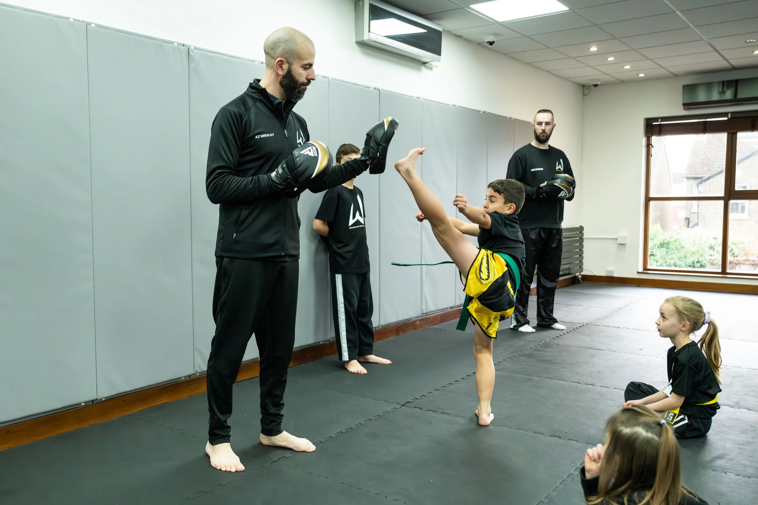 An instructor demonstrating a high kick to a young student in martial arts class, with other children sitting on the floor watching.