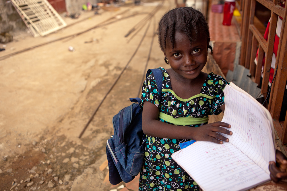 Seven-year-old Ismatu K. poses outside her home with her book bag. her older sister Mariama, also a She's the First Scholar, helps her with her homework everyday. 