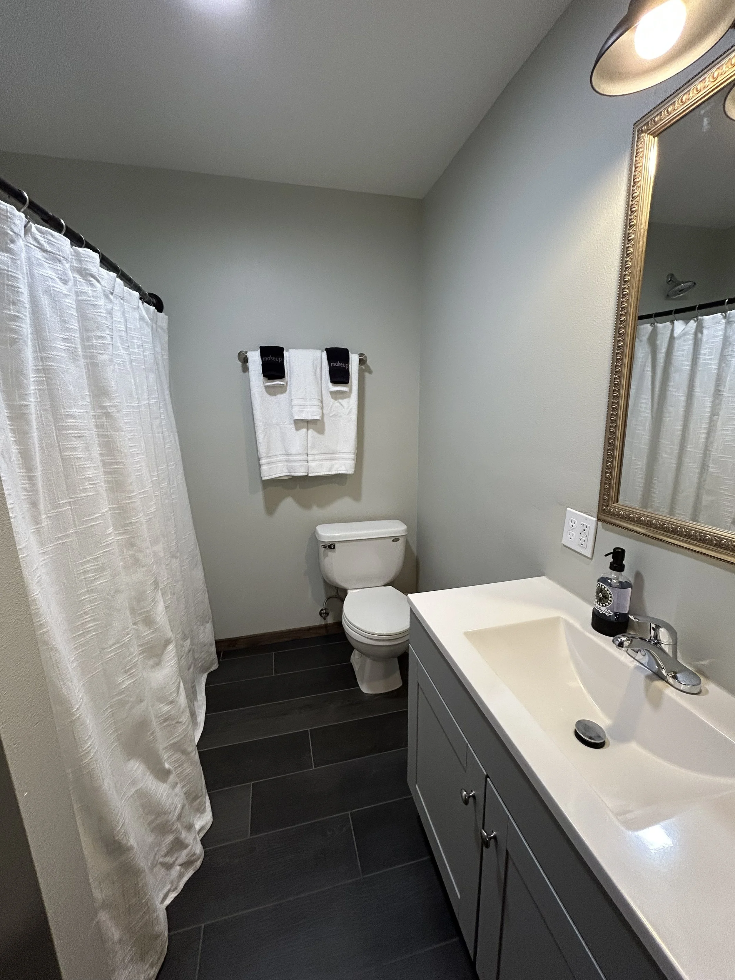 Small bathroom with a toilet, a white vanity with a sink, a mirror, a towel rack with white towels, and a white shower curtain. The floor is dark wood or tile.