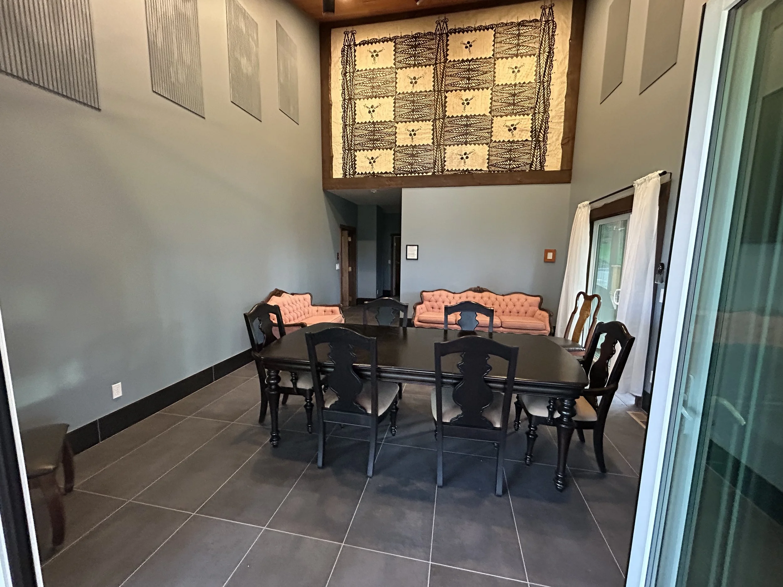 Dining room with a black wooden table, eight black chairs, two pink tufted sofas against the wall, a large decorative wall hanging, and sliding glass doors with white curtains.