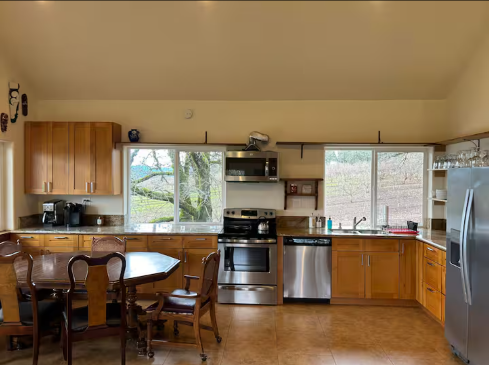 Kitchen with wooden cabinets, stainless steel appliances, a round dining table with chairs, and windows showing outdoor trees.