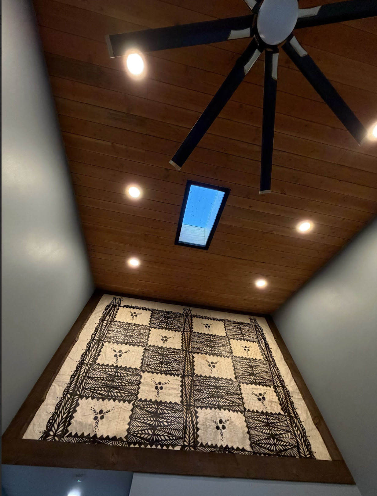 View of a ceiling with a wooden finish, ceiling lights, a black ceiling fan, and a skylight showing a blue sky.