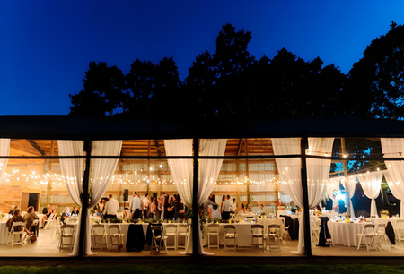 Outdoor evening reception with a glass-enclosed tent, decorated with string lights and white curtains, filled with people and banquet tables.