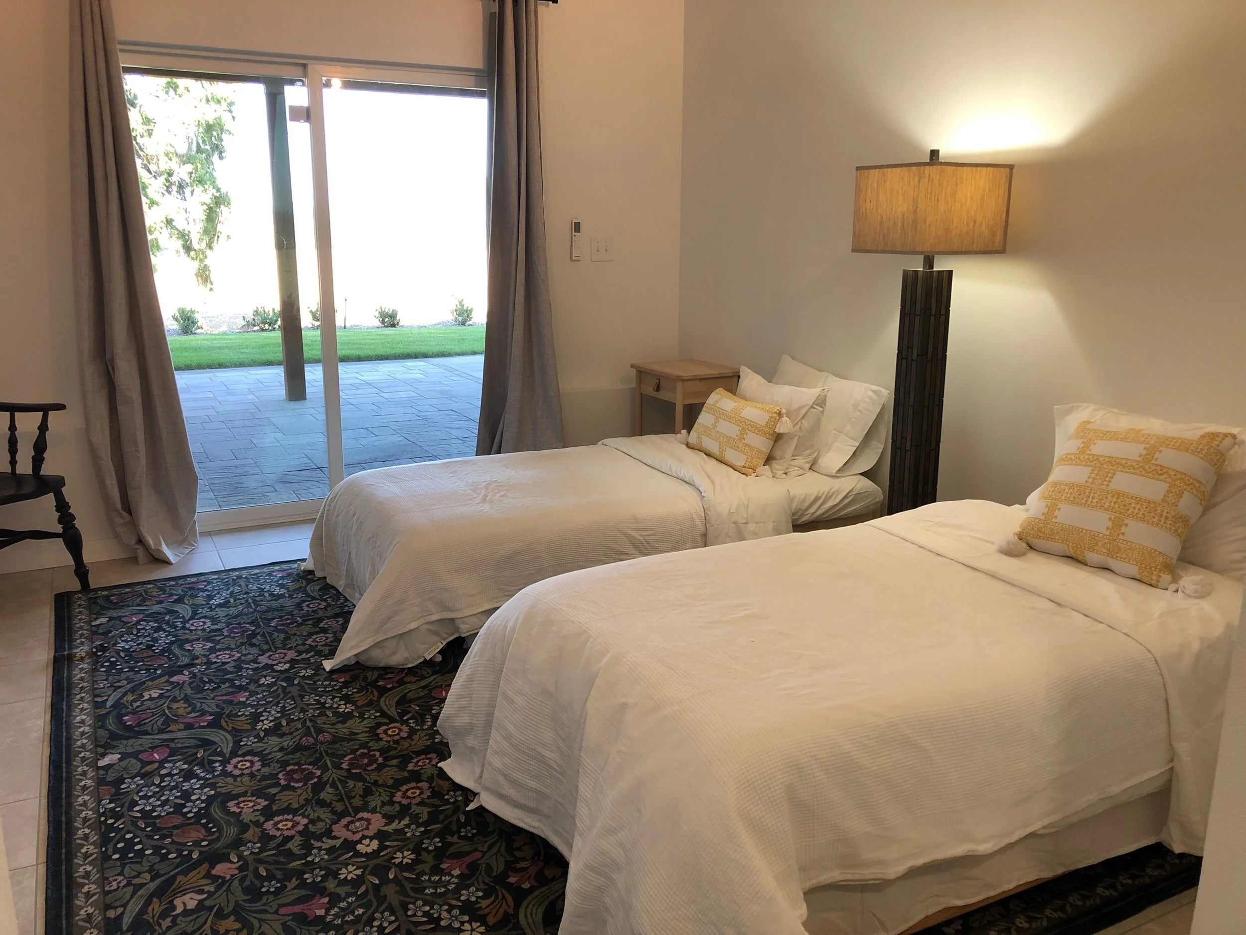 A bedroom with two white beds, decorative pillows, a nightstand, a floor lamp, a rocking chair, a rug, and sliding glass doors leading to a patio with a lawn outside.