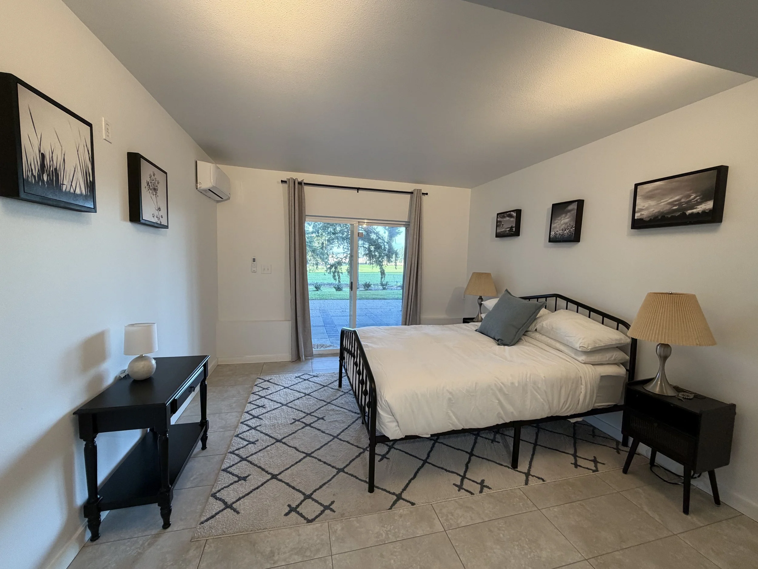 A bedroom with a black metal bed frame, white bedding, and gray pillows. Two nightstands with lamps on either side of the bed. Four framed black-and-white photos on the wall above the bed. A sliding glass door with curtains leads to an outdoor patio 