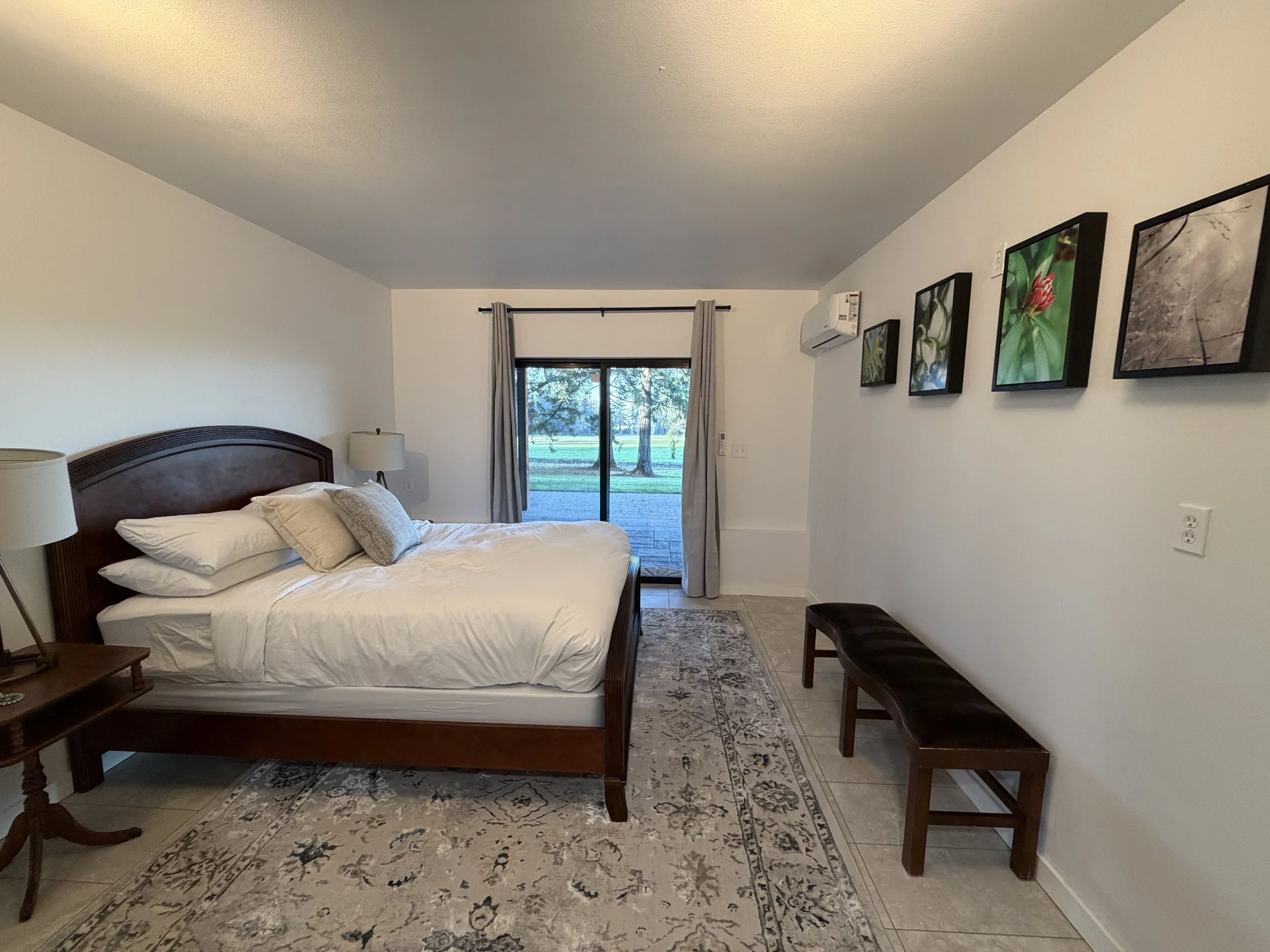 Bedroom with a bed, nightstands with lamps, an art display on the wall, a sliding glass door with curtains, and a view of trees outside.