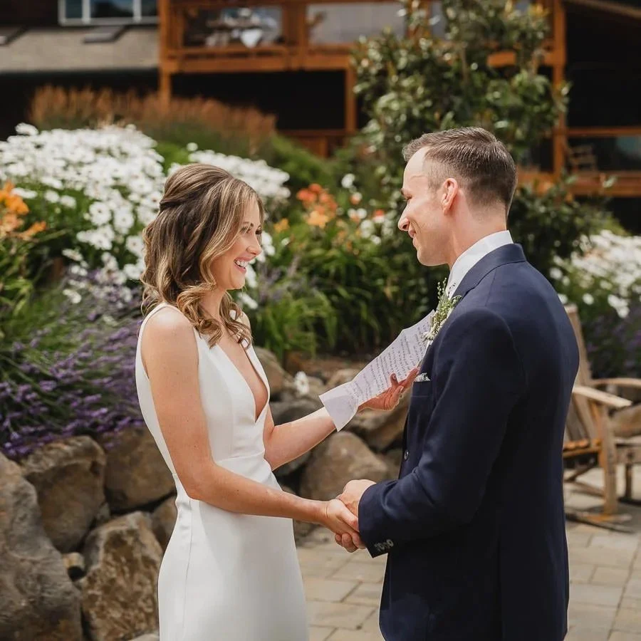 A bride and groom exchanging vows during their outdoor wedding ceremony, with flowers and a wooden house in the background.