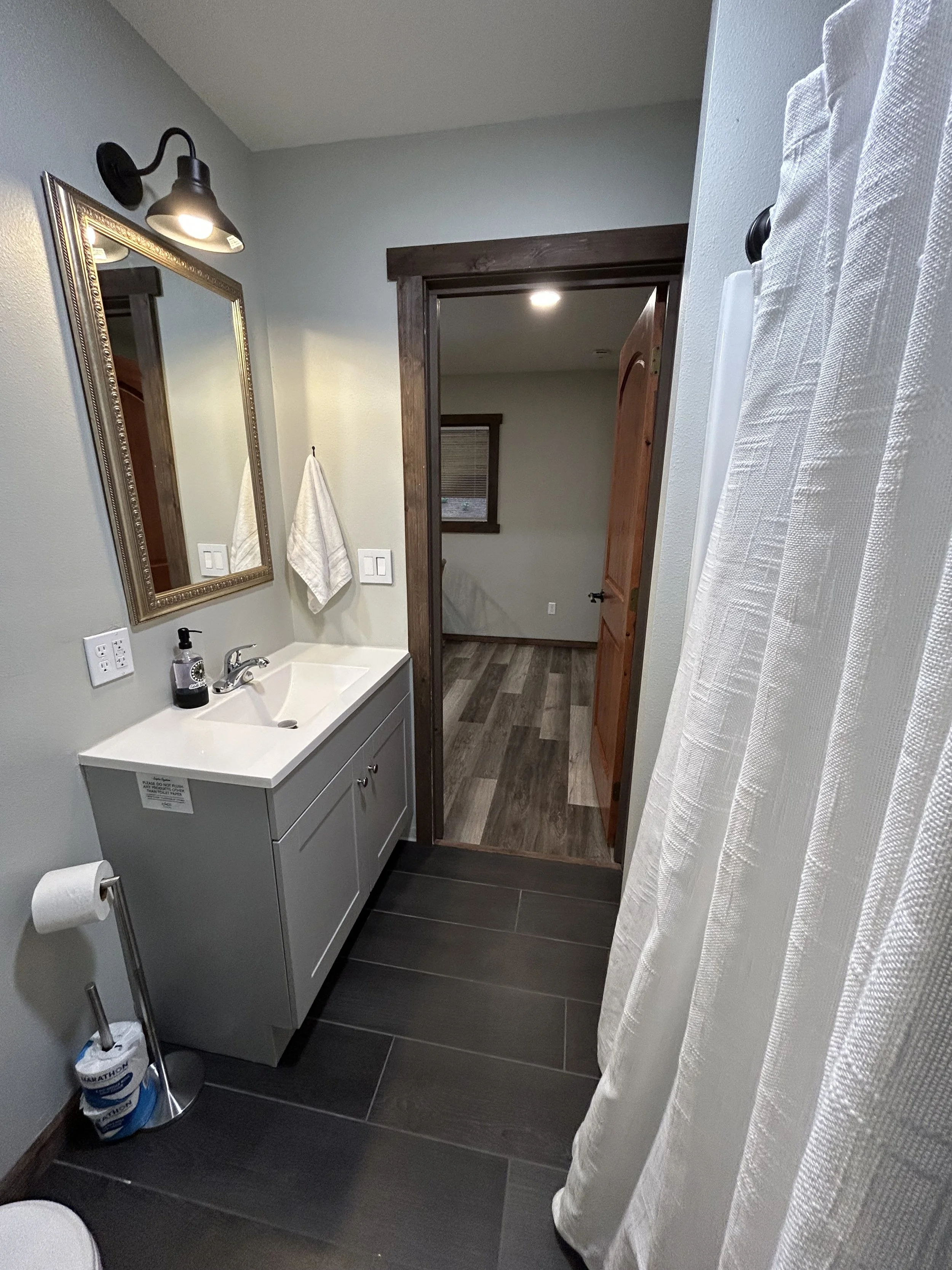 Bathroom with a small white vanity, mirror, black wall lamp, soap dispenser, hand towel, toilet paper, and a curtain on the right side, with a doorway leading to a room with wood floor and a window.