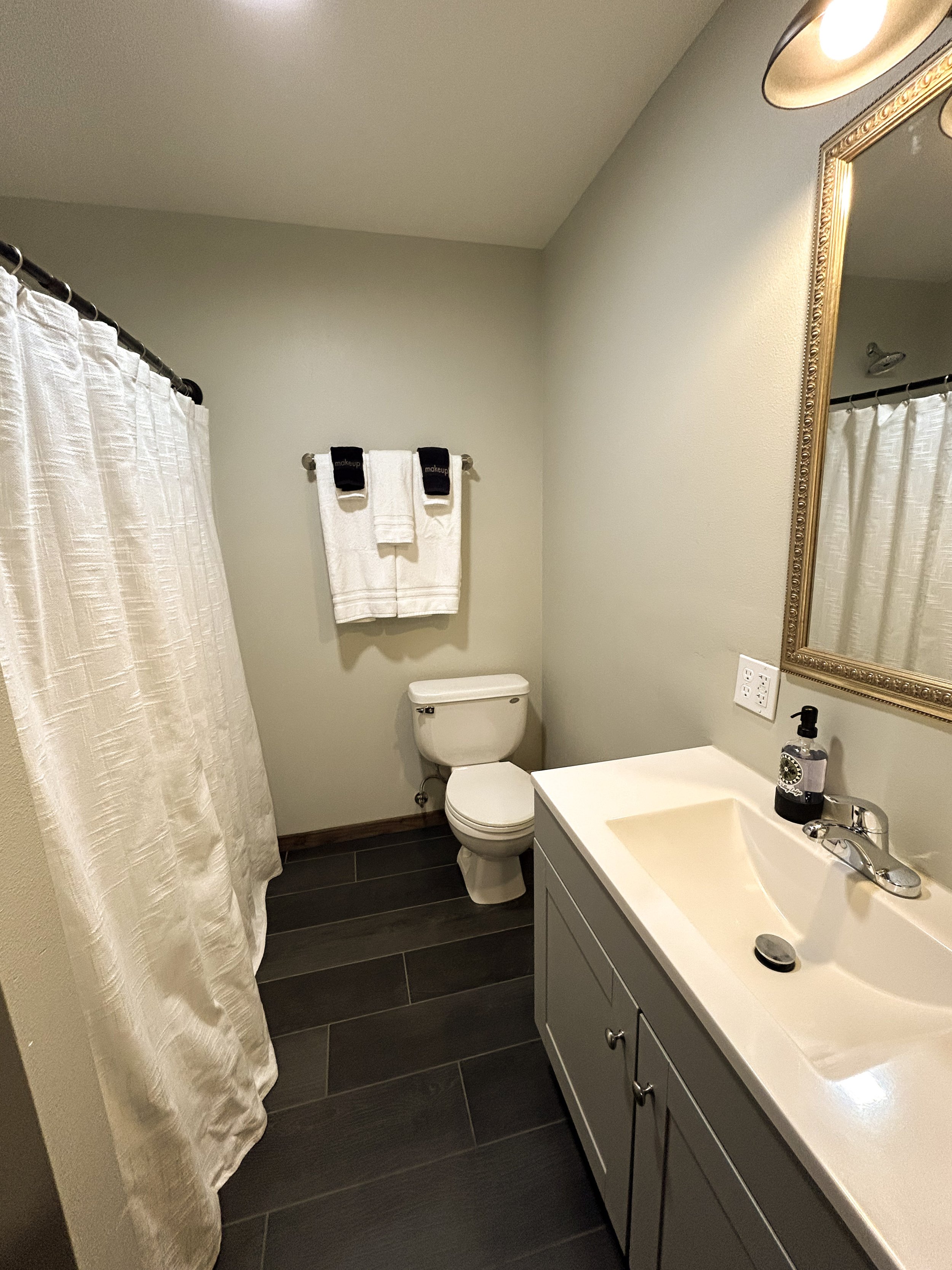 Bathroom with a white shower curtain, a toilet, a mirror with a decorative gold frame, a sink with a black soap dispenser, and white towels hanging on a towel rack.