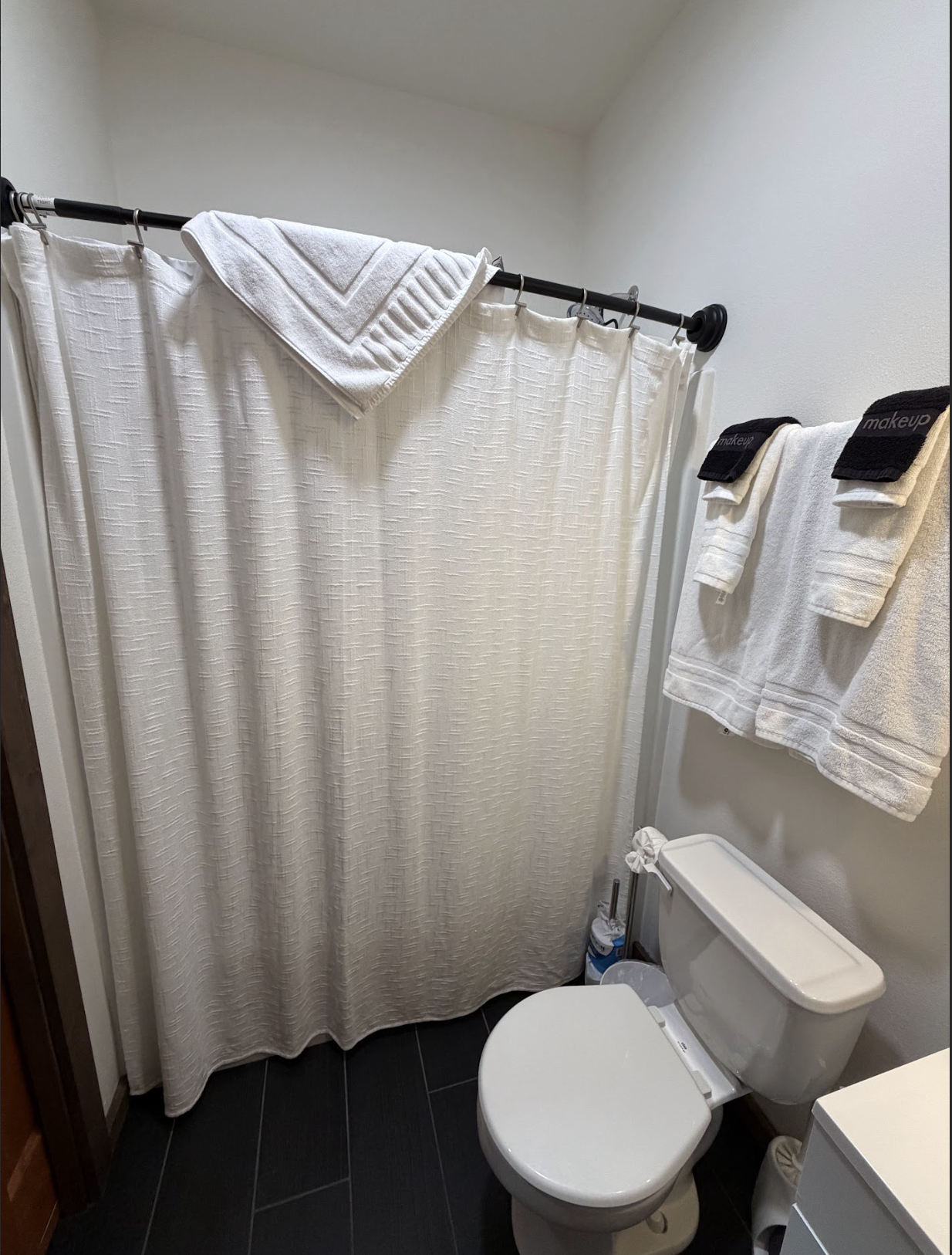 A small bathroom with a white toilet, a black shower rod with a cream shower curtain, and towel racks with white towels and black 'makeup' bands.