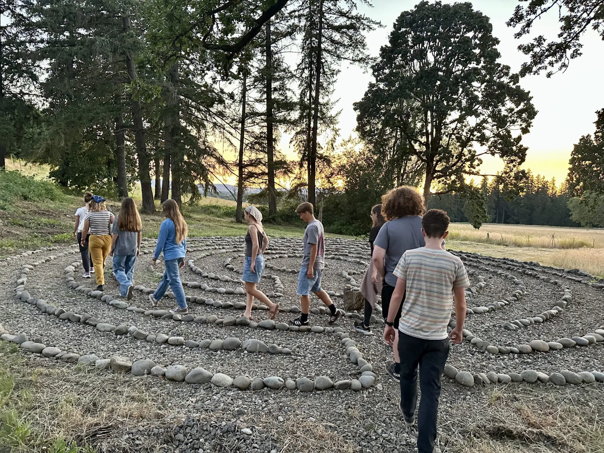 A group of people walking outdoors on a gravel path lined with rocks, surrounded by trees and greenery, with the sun setting in the background.