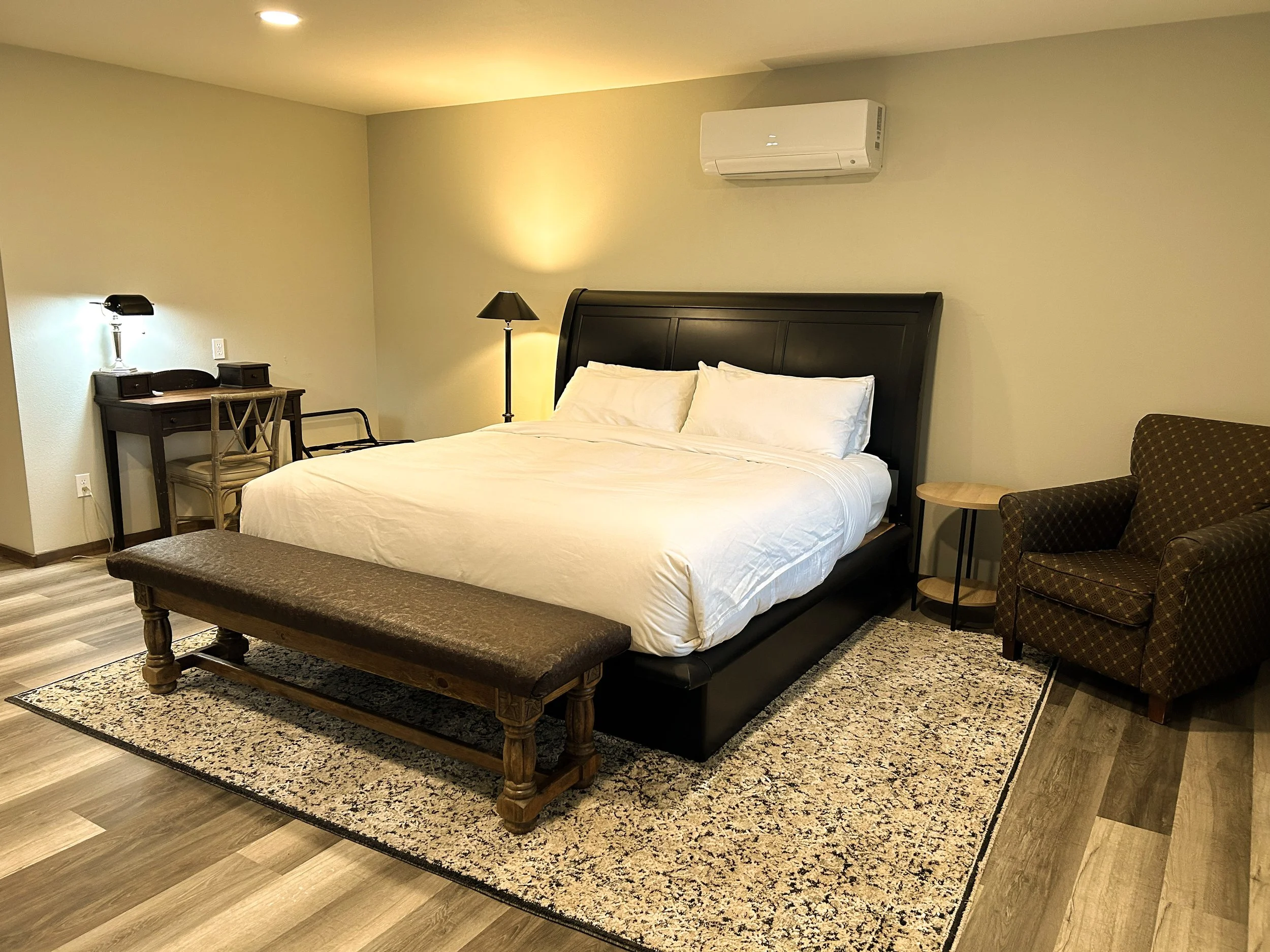 A hotel bedroom with a large bed, nightstand, desk with a lamp, armchair, and rug on hardwood floors. An air conditioning unit is mounted on the wall.