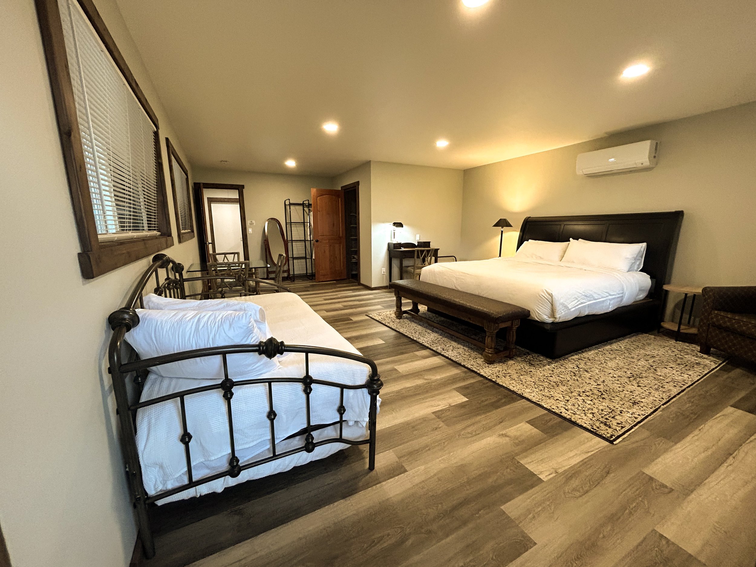 A hotel room with two beds, one with a metal frame and the other with a dark wooden frame, a small desk with a lamp, a mirror, multiple windows with blinds, and an air conditioning unit on the wall.