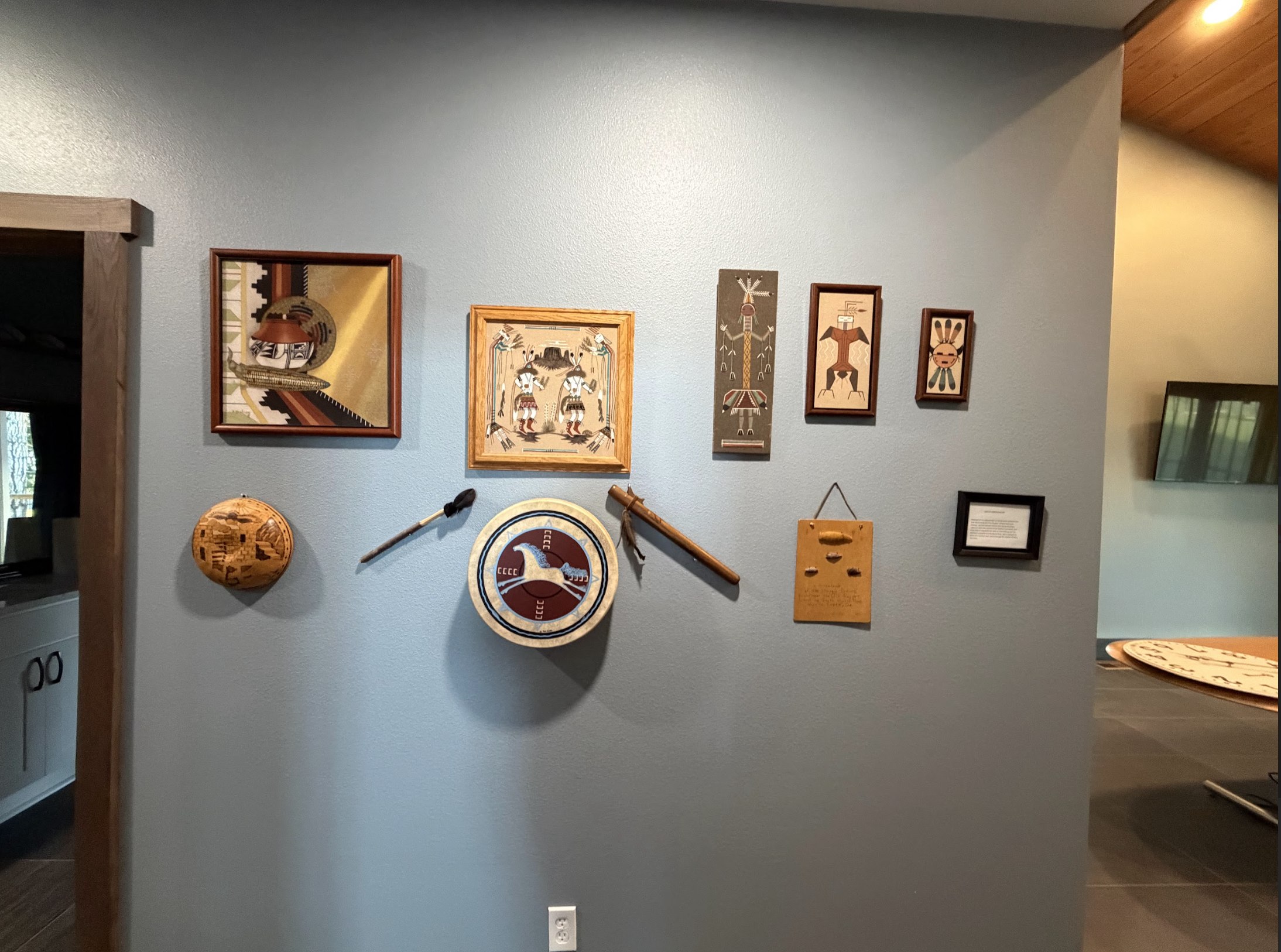 Display of Native American art and artifacts on a light blue wall, including framed paintings, woven baskets, carved wooden objects, and small sculptures.