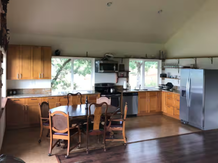 A kitchen with wooden cabinets, a dining table with six chairs, stainless steel appliances, and windows overlooking greenery.
