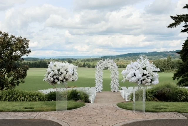 Wedding arch decorated with white flowers and two floral arrangements on tall clear stands at an outdoor ceremony site overlooking a grassy field and rolling hills.