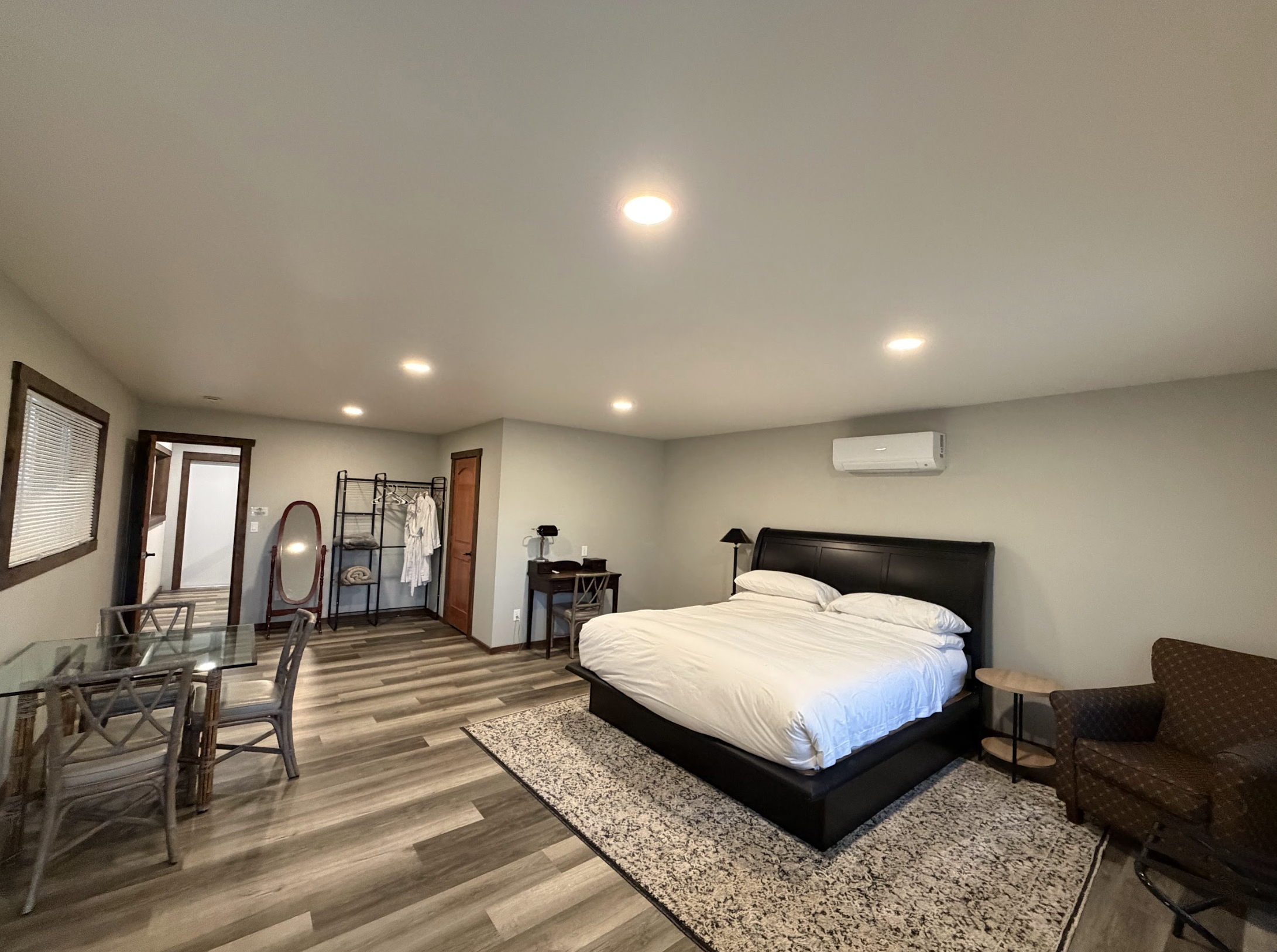 A spacious bedroom with a large bed, sitting on a patterned rug, two side tables, a desk with a chair, a clothes rack, a mirror, and a sitting armchair.